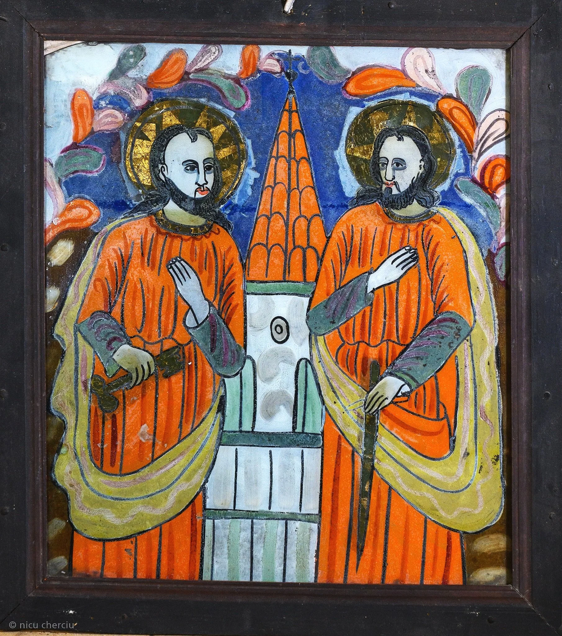 A colorful religious icon painting depicting two saints in orange robes with halos, holding swords, standing in front of a church with a tall spire, with clouds and a blue sky in the background.