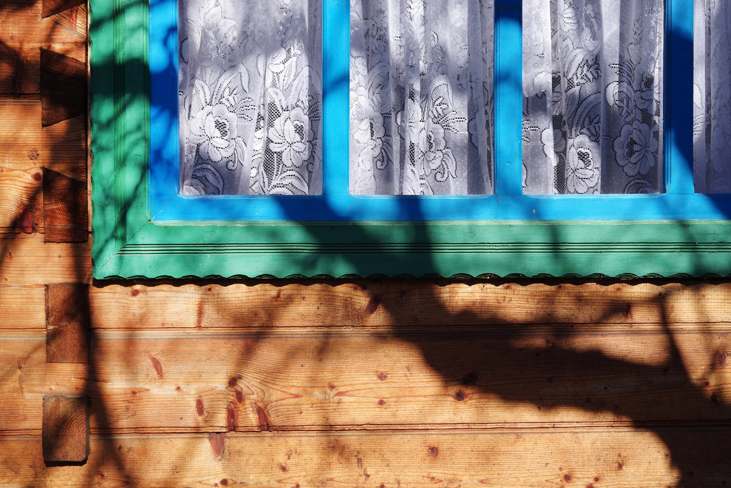 A close-up of a wooden house wall with a window. The window has a blue frame and white lace curtains, with shadows cast across the wall.