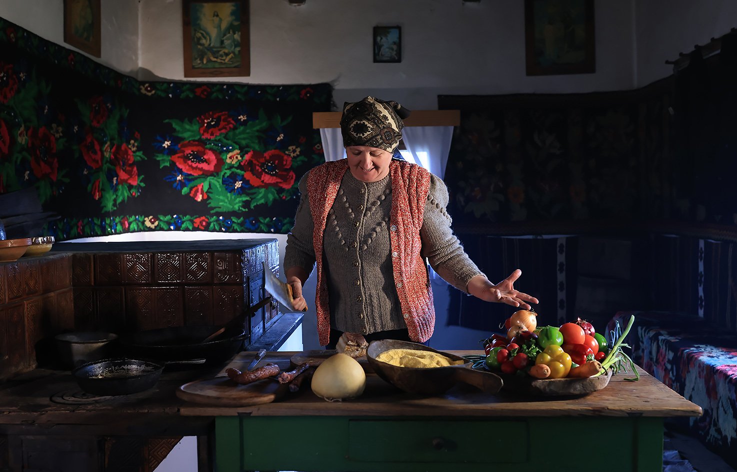 A woman in traditional clothing, including a headscarf and knit sweater, stands in a rustic kitchen with a wooden table, surrounded by vegetables, herbs, and kitchen utensils, in a cozy interior decorated with floral and tapestry wall hangings.