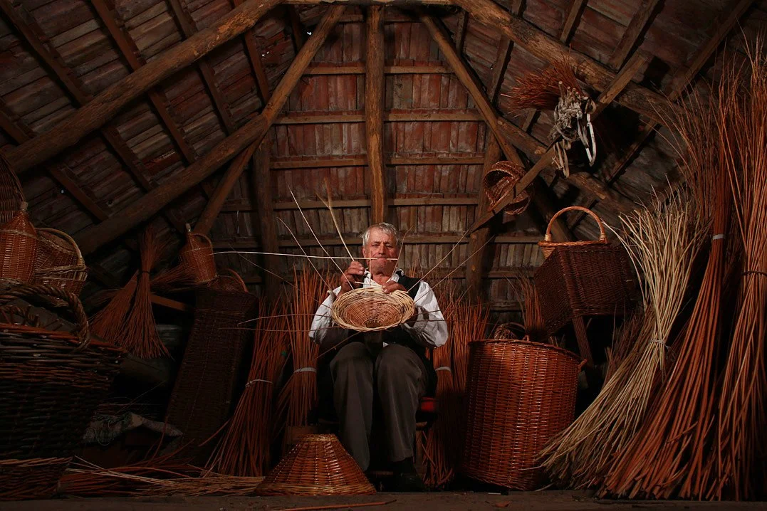 An elderly man weaving a basket inside a rustic wooden attic filled with other woven baskets and natural materials.