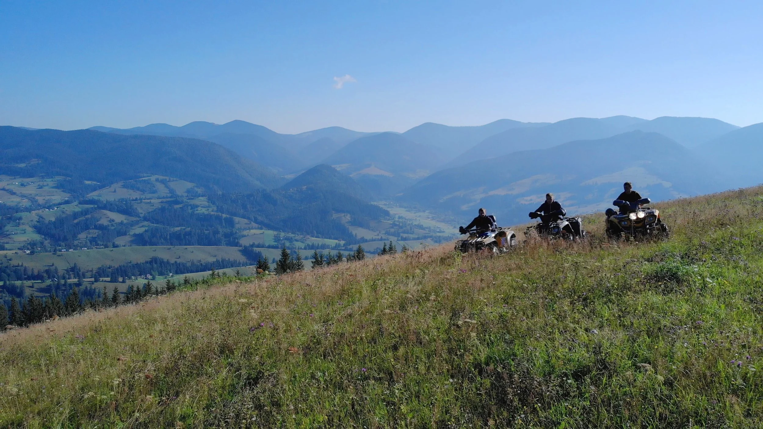Three people riding all-terrain vehicles (ATVs) on a grassy hillside with mountains in the background under a clear blue sky.