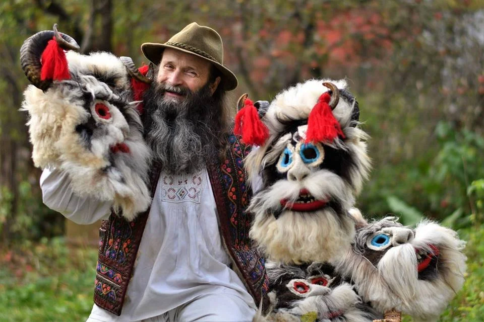 A man with a long beard and hat smiling while holding two large colorful puppet masks with horns, big eyes, and furry textures outdoors.