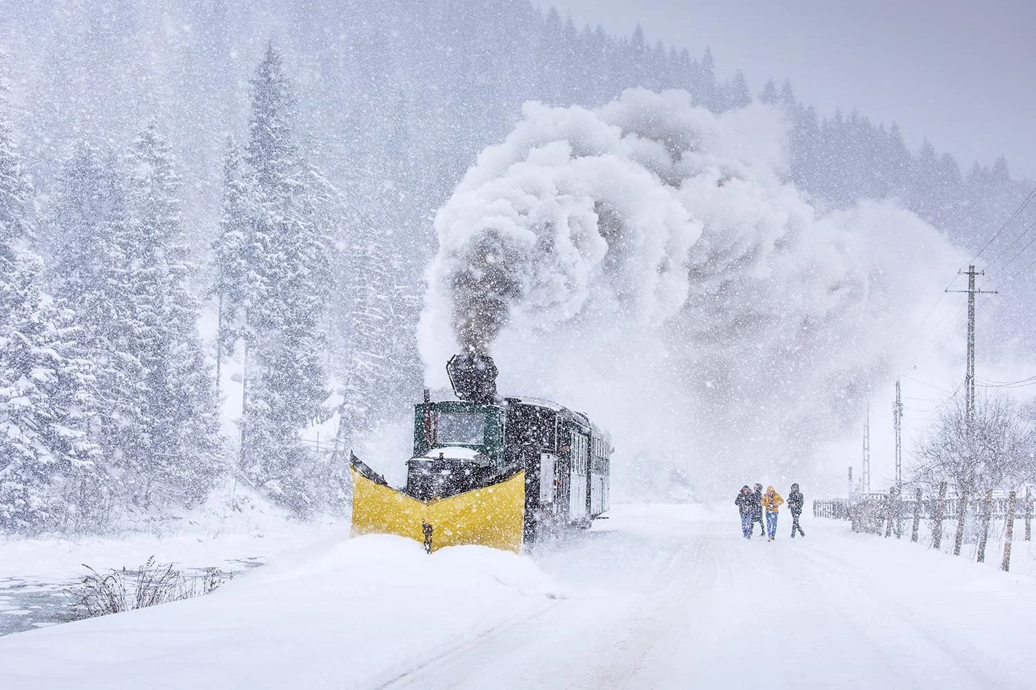 A diesel train with a yellow snowplow on the front is clearing snow on a snowy railway track in a winter landscape. There are three people walking on the right side, and snow-covered trees in the background.