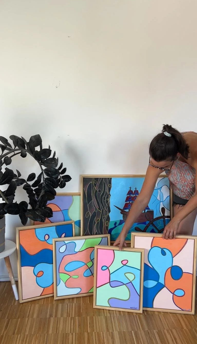 Eine Person sortiert bunte abstrakte Gemälde, die gegen eine weiße Wand und Holzfußboden gelehnt sind, daneben steht eine große schwarze Topfpflanze.