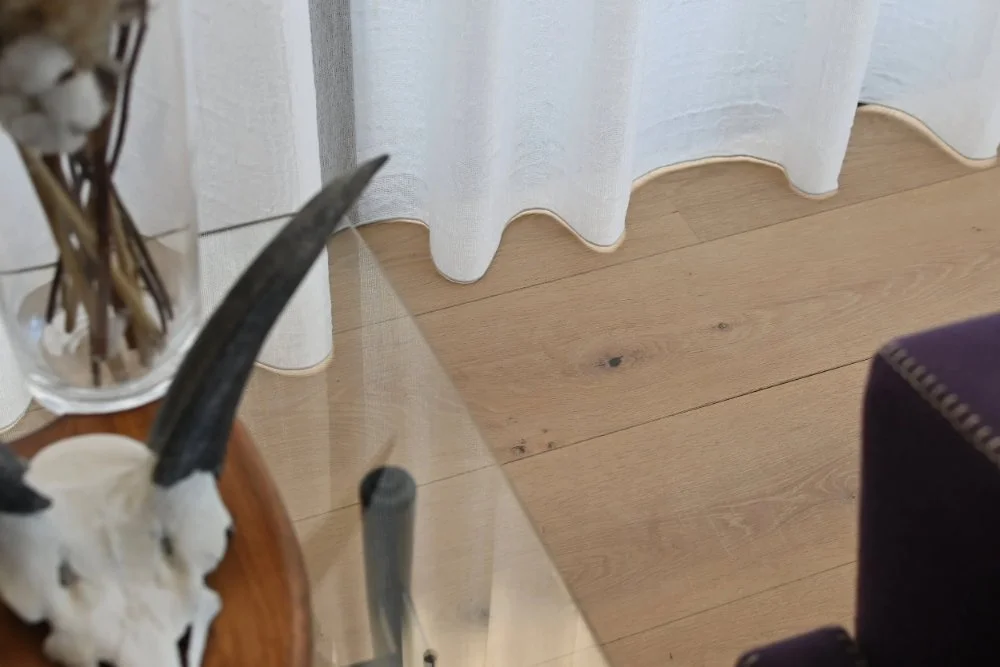 Close-up van een witte lichte vitrage gordijn dat over de houten vloer hangt. In de linkerhoek staat een glas met wat drogen bloemstengels.