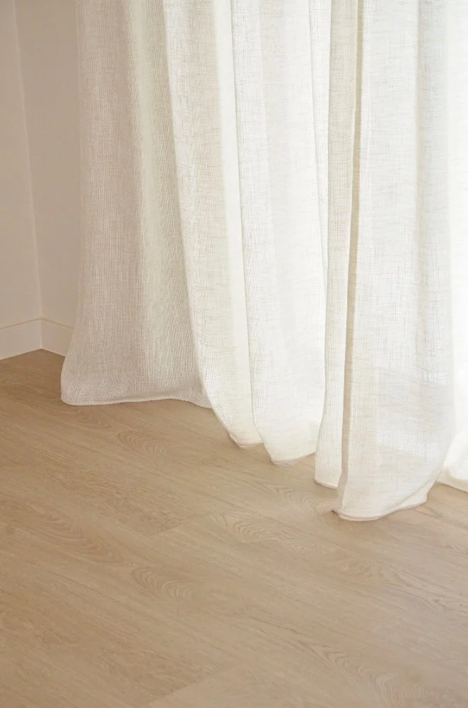 Lichtkleurige gordijnen hangen voor een raam tegen een lichte houten vloer.