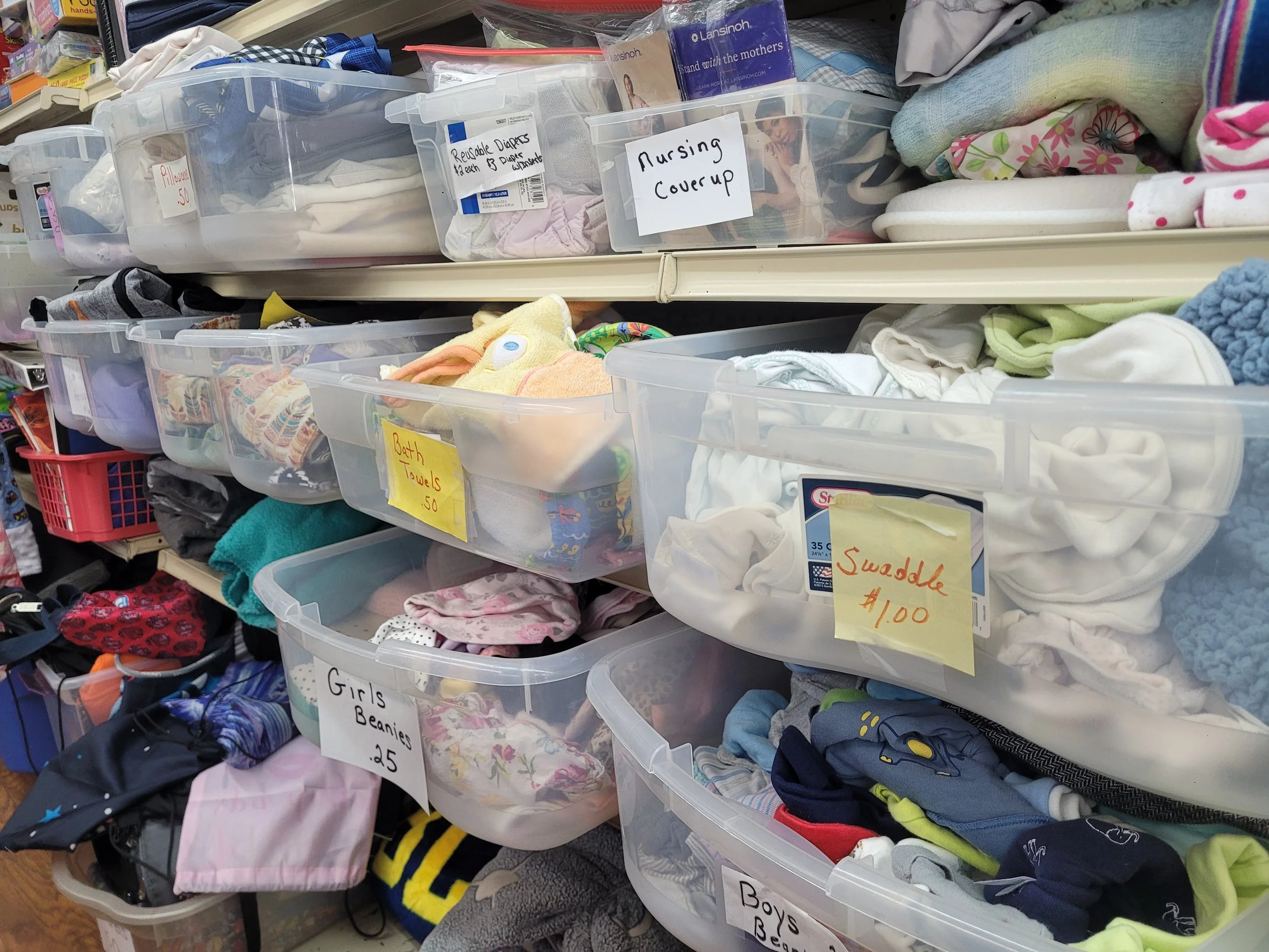 Clear plastic bins filled with children's clothing, labeled with signs indicating items for sale such as "Girls Beanies 25," "Bath Towels 50," "Swaddle 1.00," and "Nursing Cover Up." Sorted clothing items are visible inside the bins, with some folded