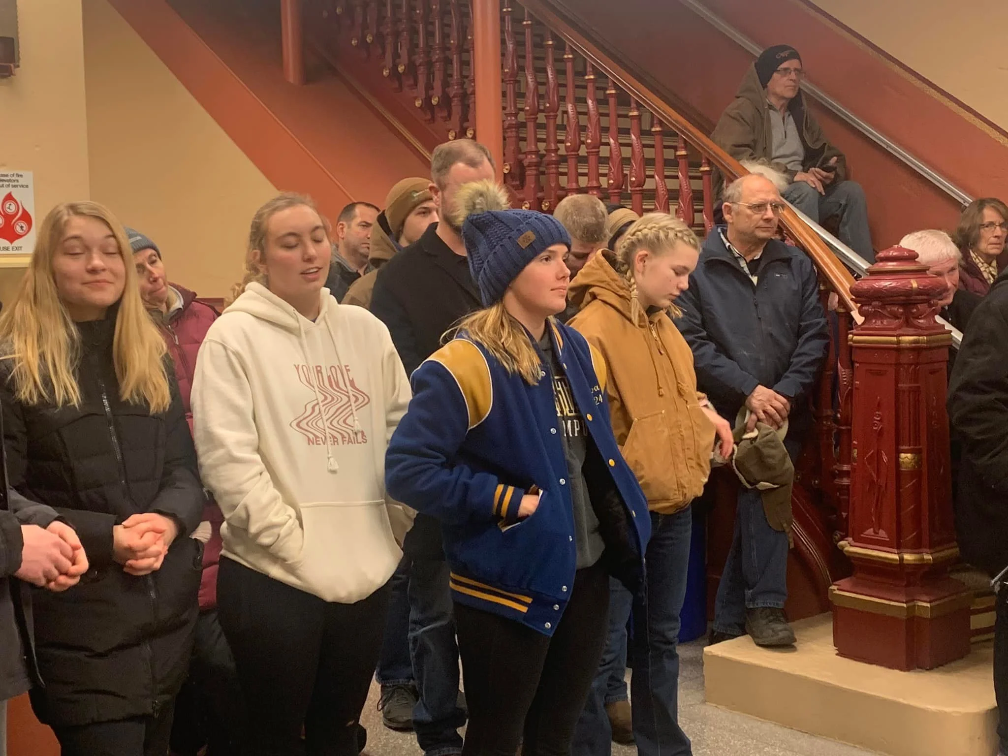Group of people standing and praying inside a building, some with eyes closed, others with hands clasped or in pockets, near a staircase with red wooden railings.