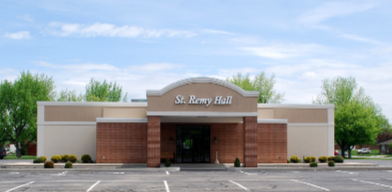 Front view of St. Remy Hall with a brick entrance and a sign on top. Green trees and a parking lot are visible in the background.
