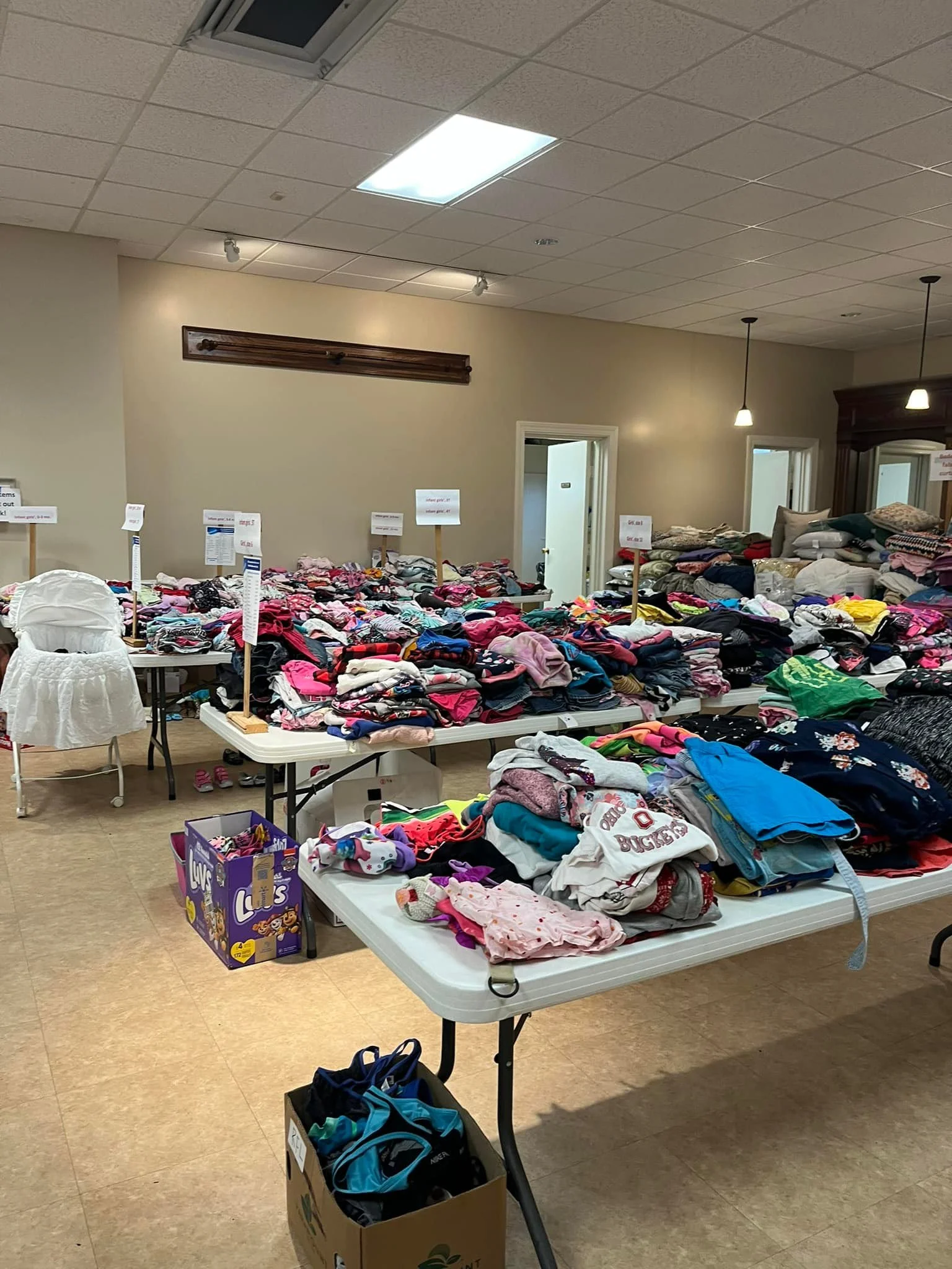 Multiple tables filled with assorted clothing, including children's clothes, with signs indicating different categories, in a room with beige walls and a ceiling with fluorescent lights.