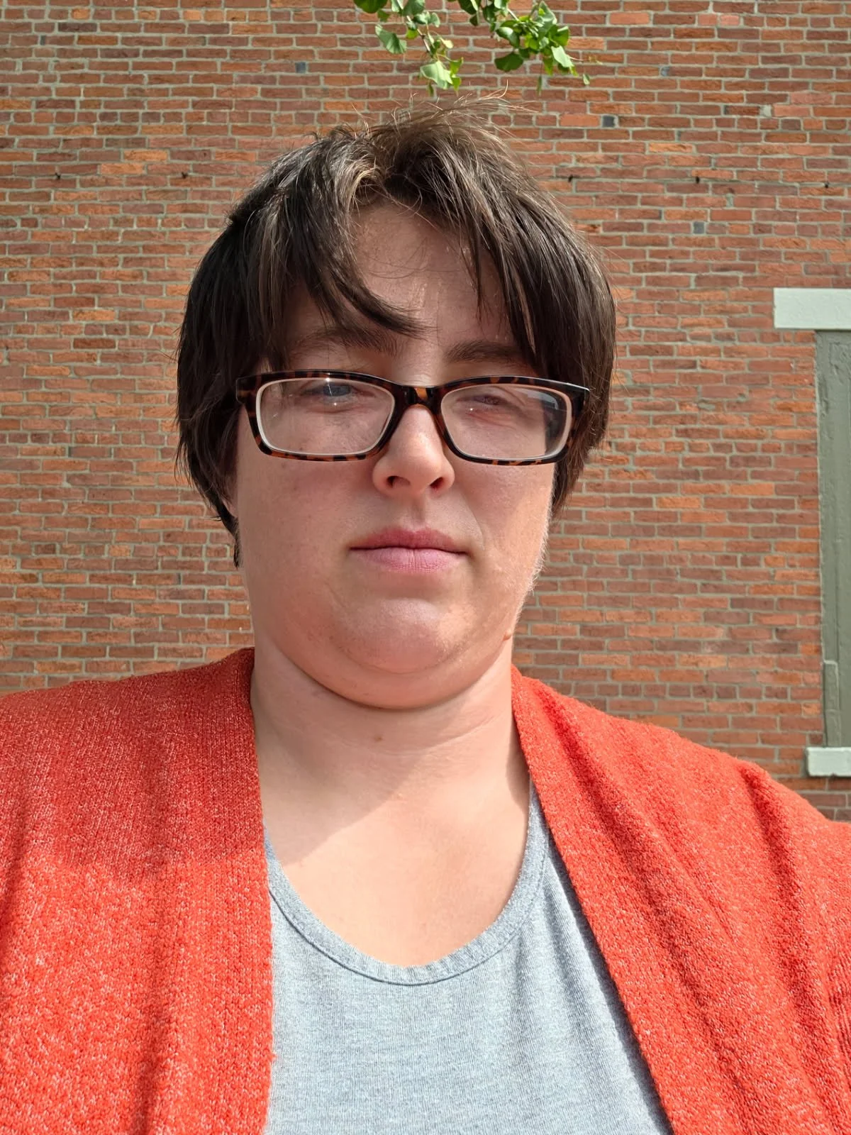 A person with short brown hair, wearing glasses and a red cardigan, standing outdoors in front of a brick wall and some green leaves