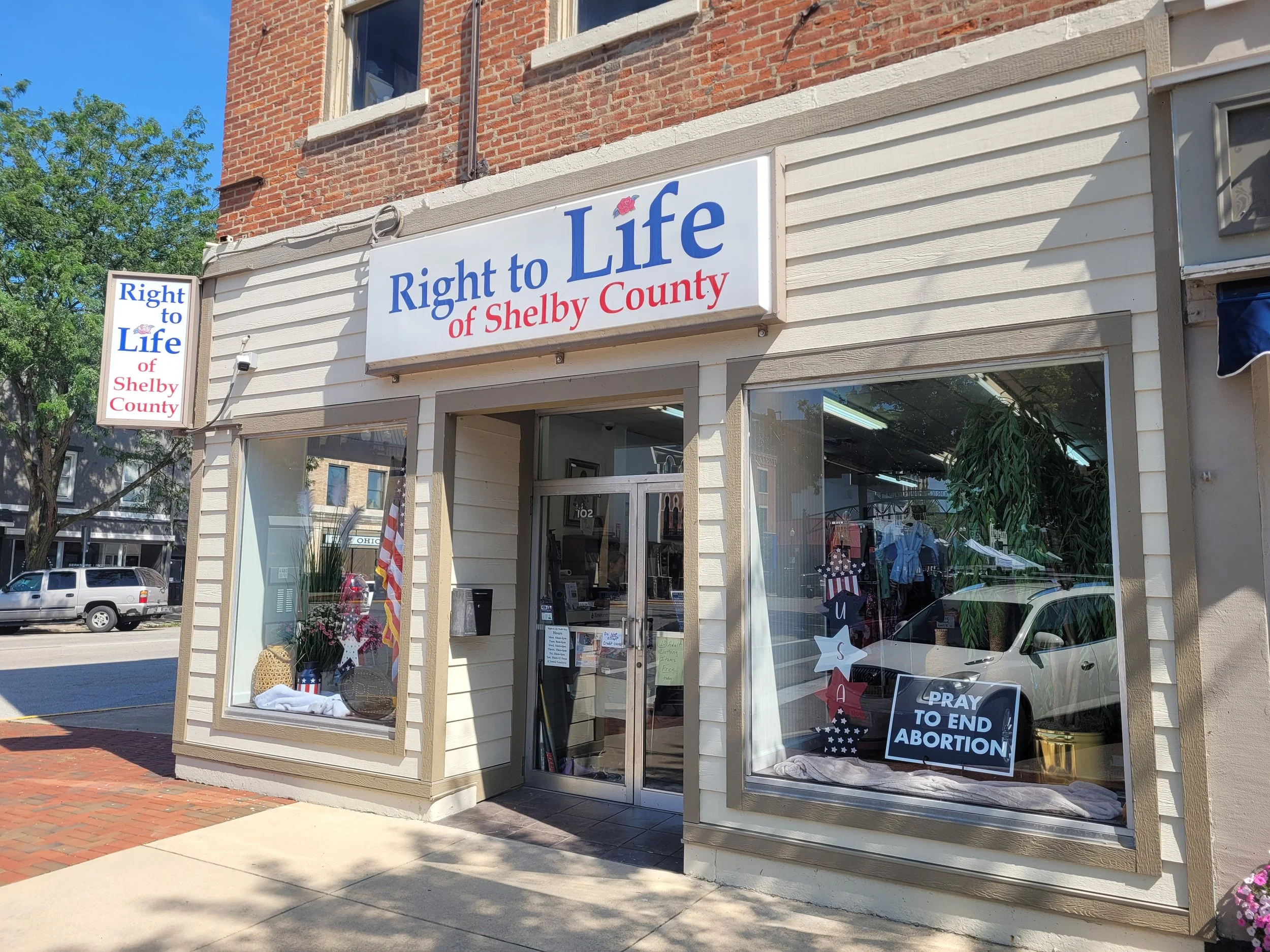  storefront of a pro-life organization named "Right to Life of Shelby County" with signs in the window encouraging prayer to end abortion, American flags, and floral arrangements inside.