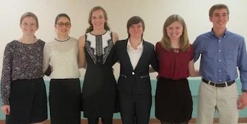Six young adults standing together in a row, dressed in casual to semi-formal attire, smiling for the photo.