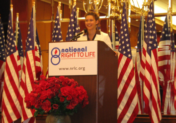 Shelby County Right to Life Oratory Contest; A woman speaking at a podium with a 'National Right to Life' sign, surrounded by American flags and red flowers.