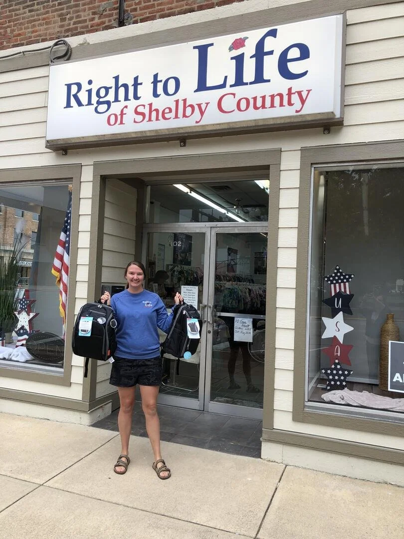 A young woman standing outside a store called 'Right to Life of Shelby County,' holding two breast pump bags and smiling.