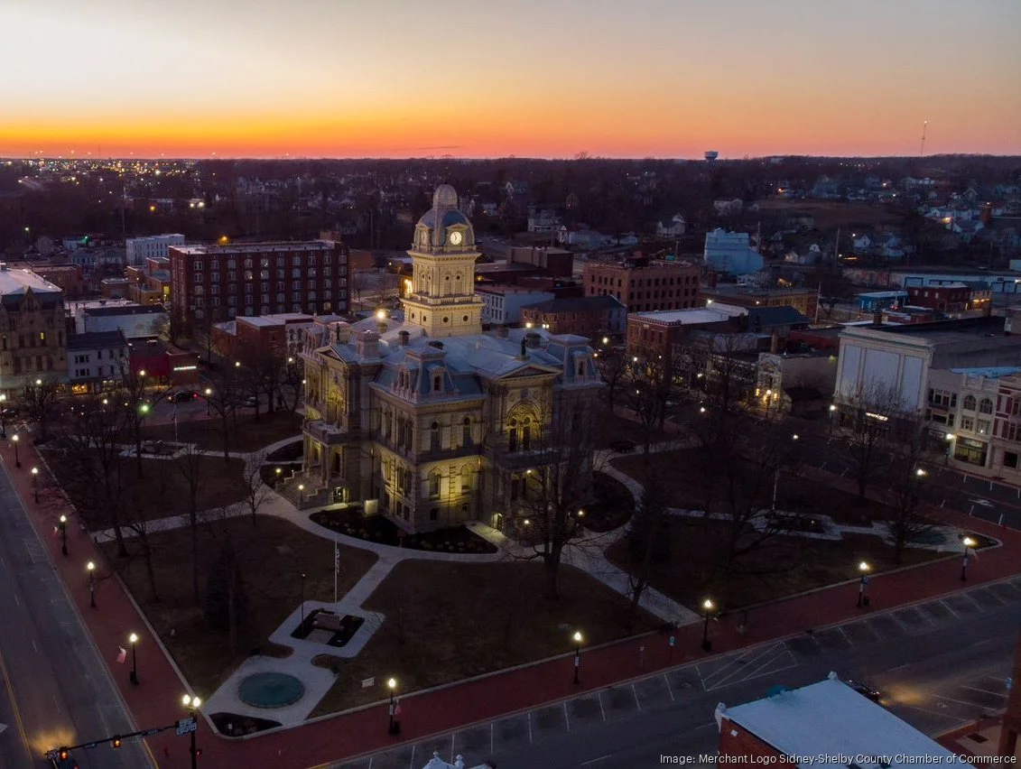 A historic courthouse in a small town at sunset, with a park, streetlights, and surrounding buildings.
