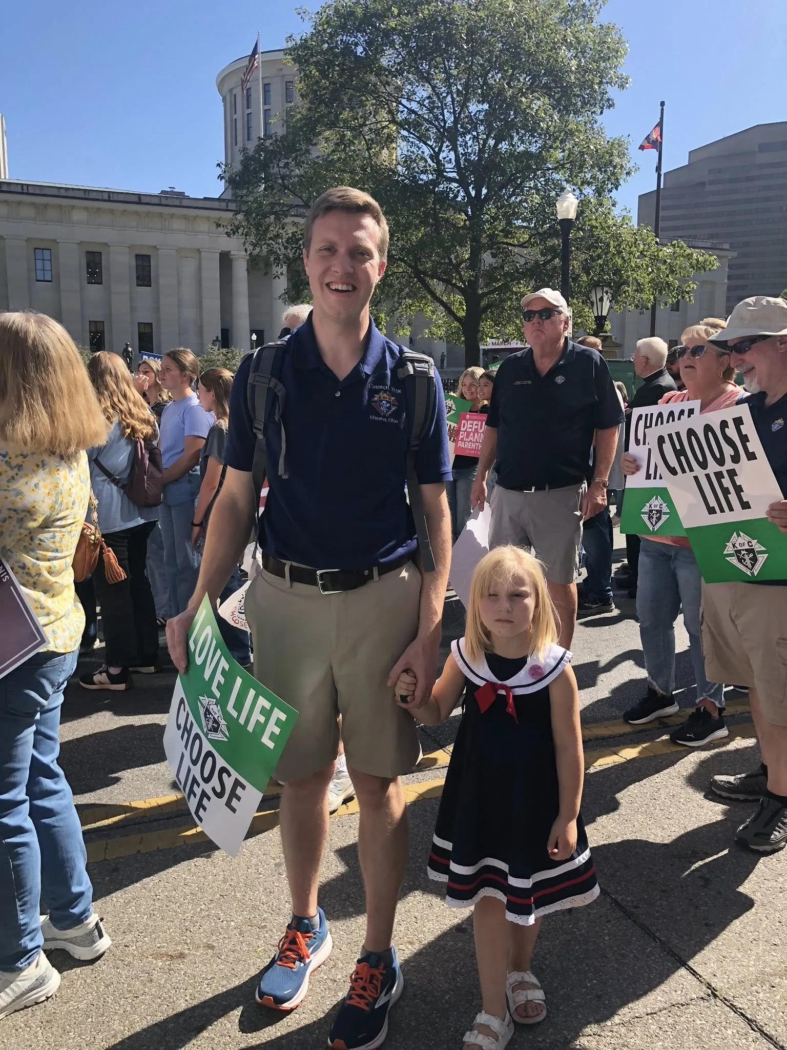 A man and a young girl holding hands at a protest rally, with people holding signs that say 'LOVE LIFE' and 'CHOOSE LIFE', in front of a government building on a sunny day.