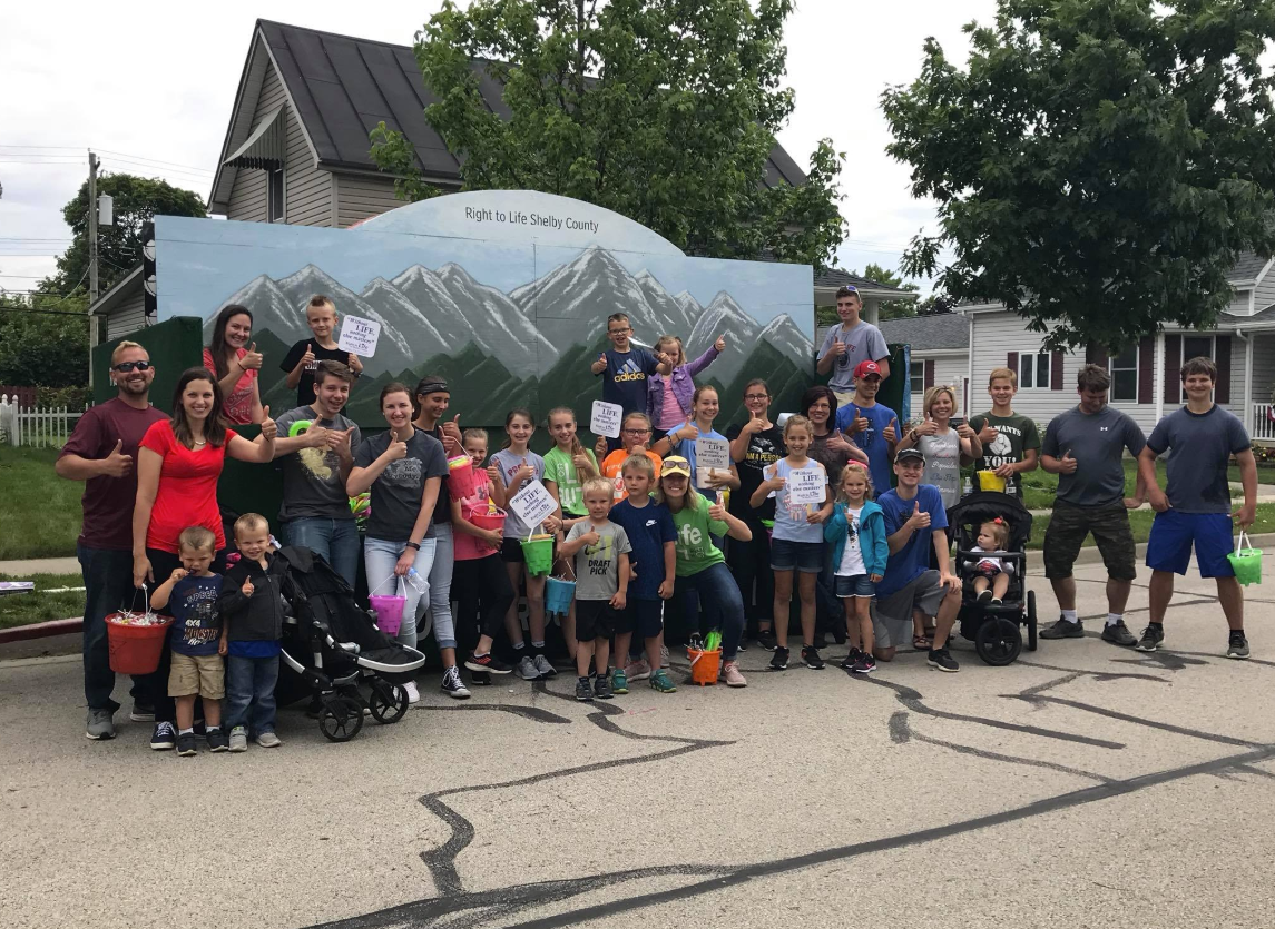 Group of children and adults outdoors celebrating, holding signs and buckets of candy, in front of a mural of mountains with a sign that says "Right to Life Shelby County."