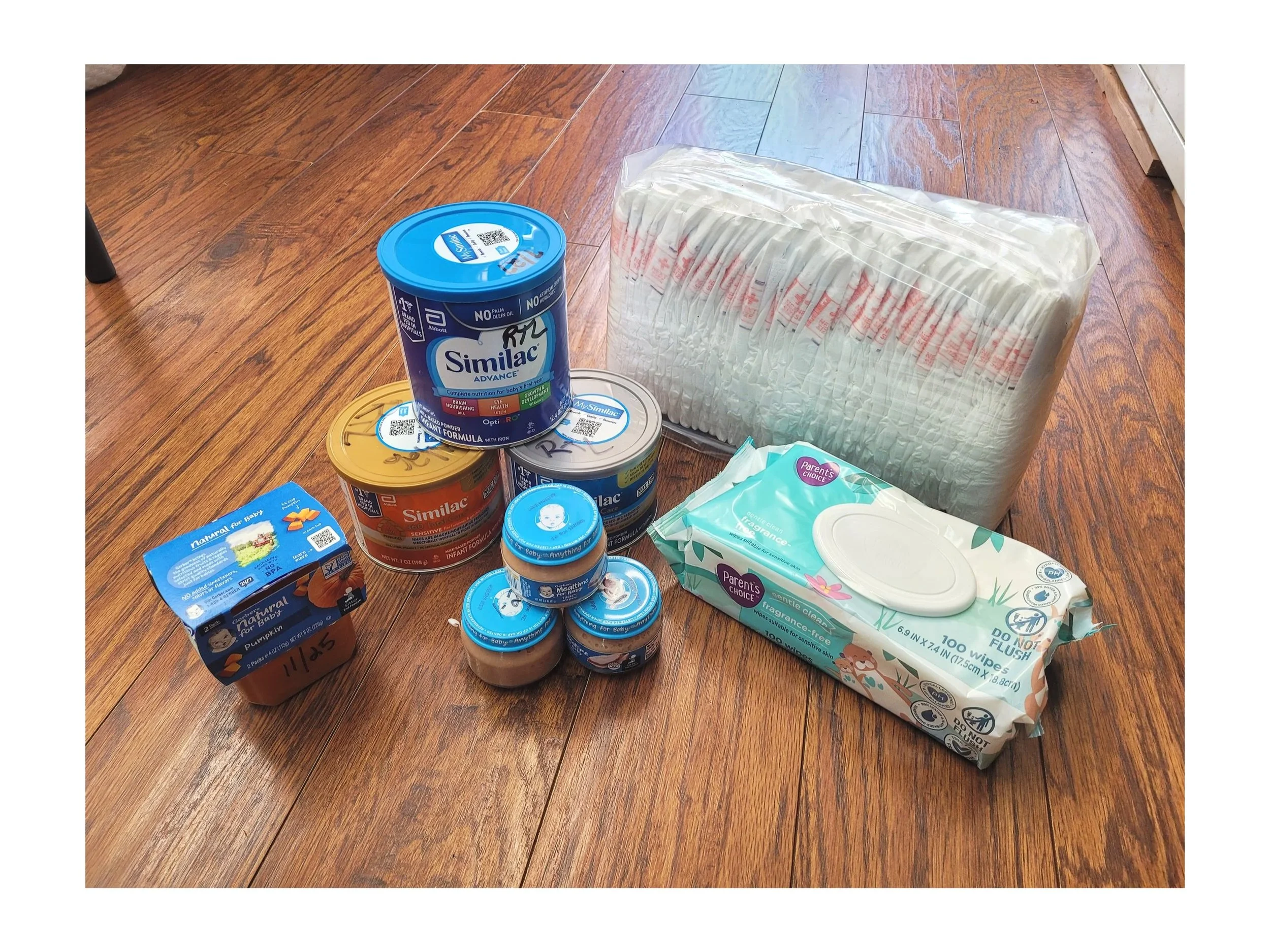 Assorted baby supplies on a wooden floor, including cans of formula, a pack of diapers, baby wipes, diaper rash ointment, and a box of baby food.