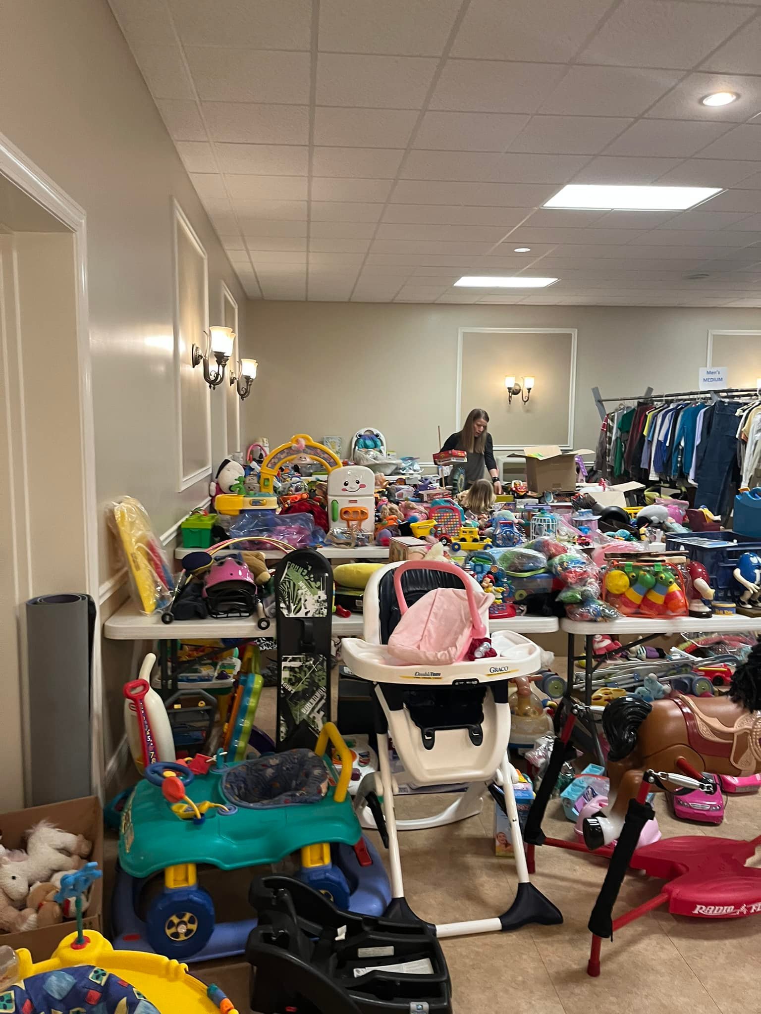 Sale of children's toys, clothes, and accessories in a large indoor space, with tables filled with colorful toys, a high chair, ride-on toys, and a clothing rack in the background.