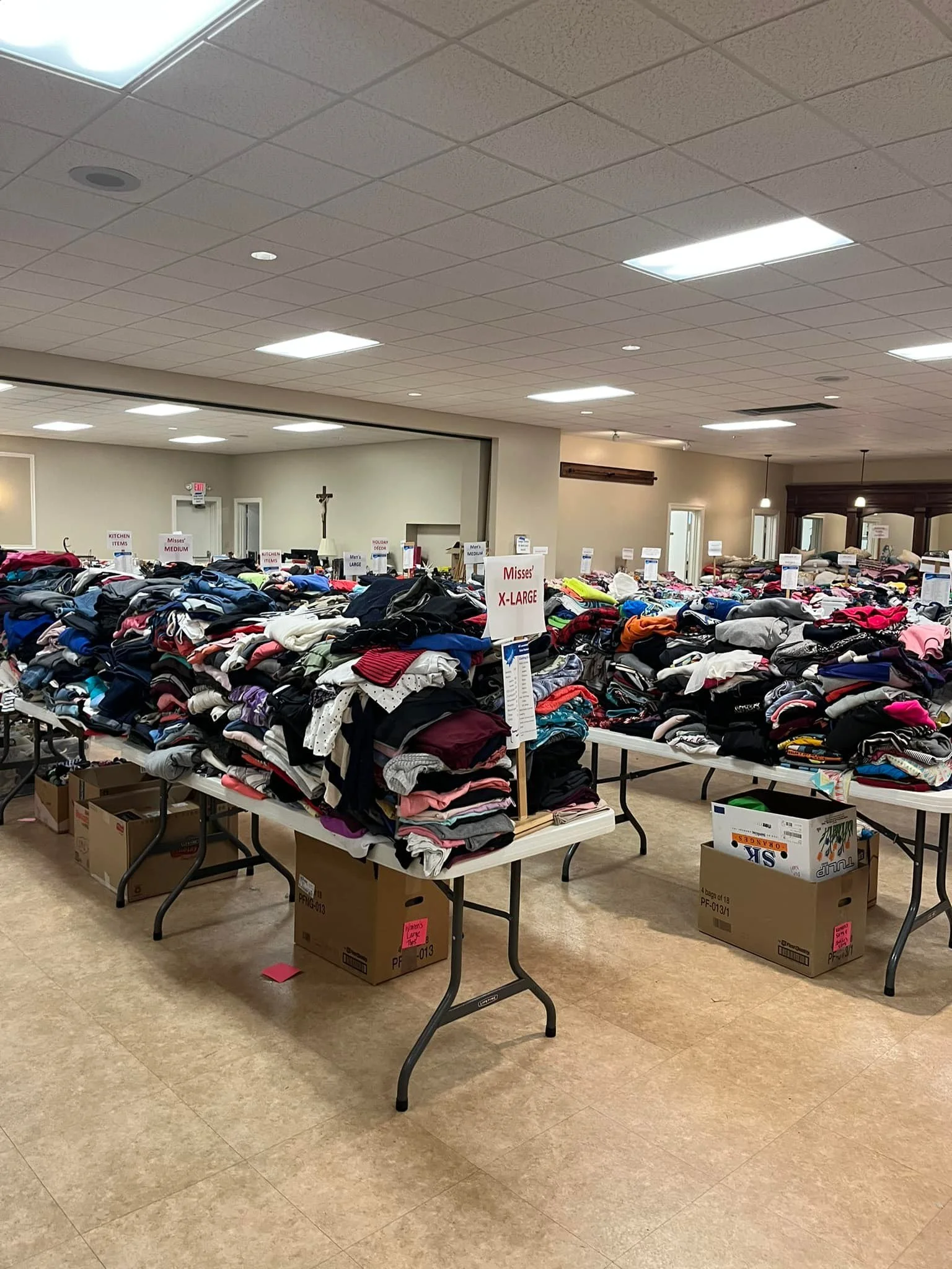 Tables filled with folded clothes at a thrift or donation center, with cardboard boxes underneath for storage, and signs indicating sizes and categories.