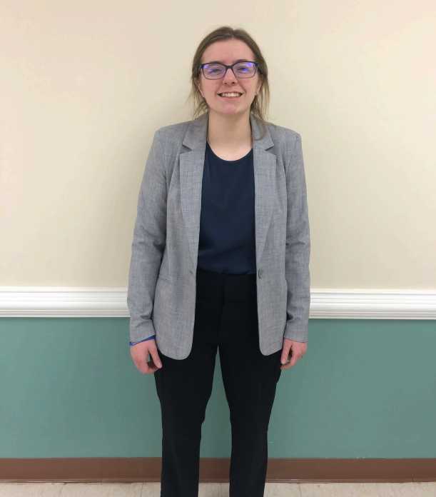 A woman standing in front of a wall with a chair rail, wearing glasses, a gray blazer, a navy blue shirt, black pants, smiling with her hands relaxed at her sides.