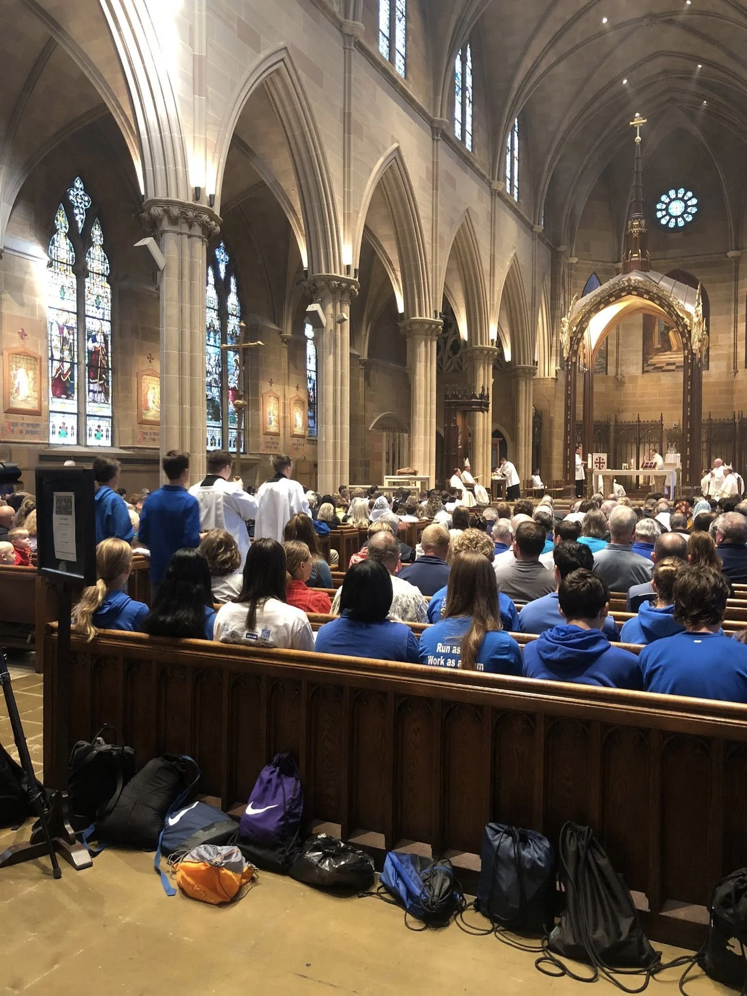 A church interior during a service with congregants seated and altar at the front. Many backpacks are on the floor at the back of the church.