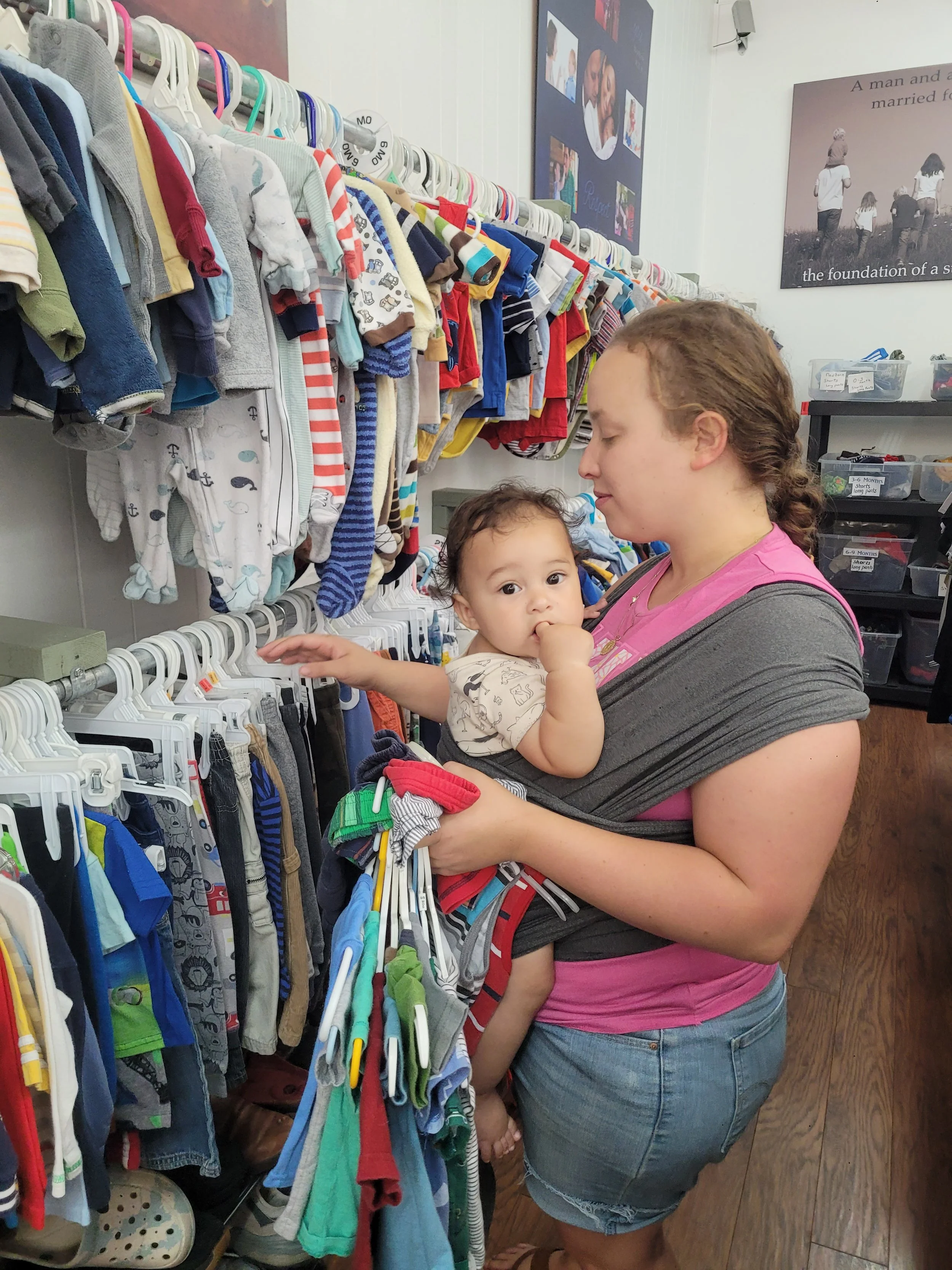 Shelby County Right to Life thrift store in Sidney, OH; A woman holding a young child in a clothing store, looking at baby clothes on hangers. The store has racks of children's clothes and photos on the wall.