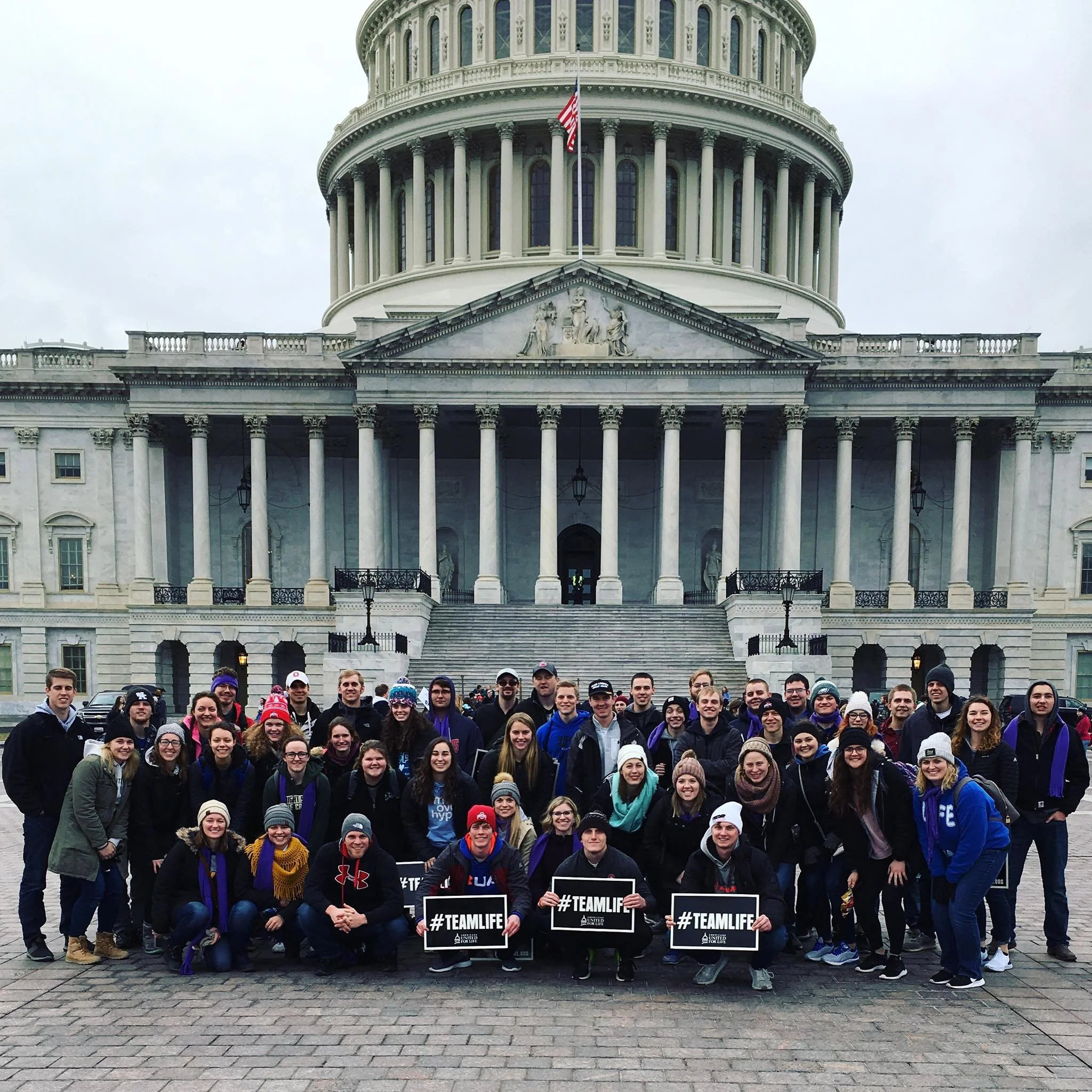 A large group of people posing for a photo in front of the United States Capitol building, some holding signs that say #TEAMLIFE, dressed warmly for cold weather. Shelby County Right to Life at the National Right to Life March in Washington DC.