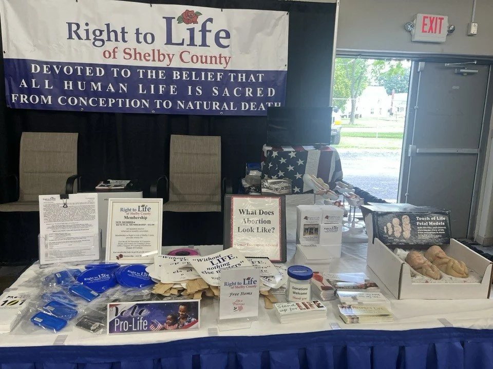 Table display by Right to Life of Shelby County with informational flyers, blue wristbands, pamphlets, and a box of plaster femur models. A large banner in the background reads "Right to Life of Shelby County, Devoted to the Belief That All Human Lif
