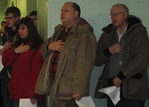 Group of people standing indoors, with some holding their hands over their hearts, possibly during a national anthem or ceremony.