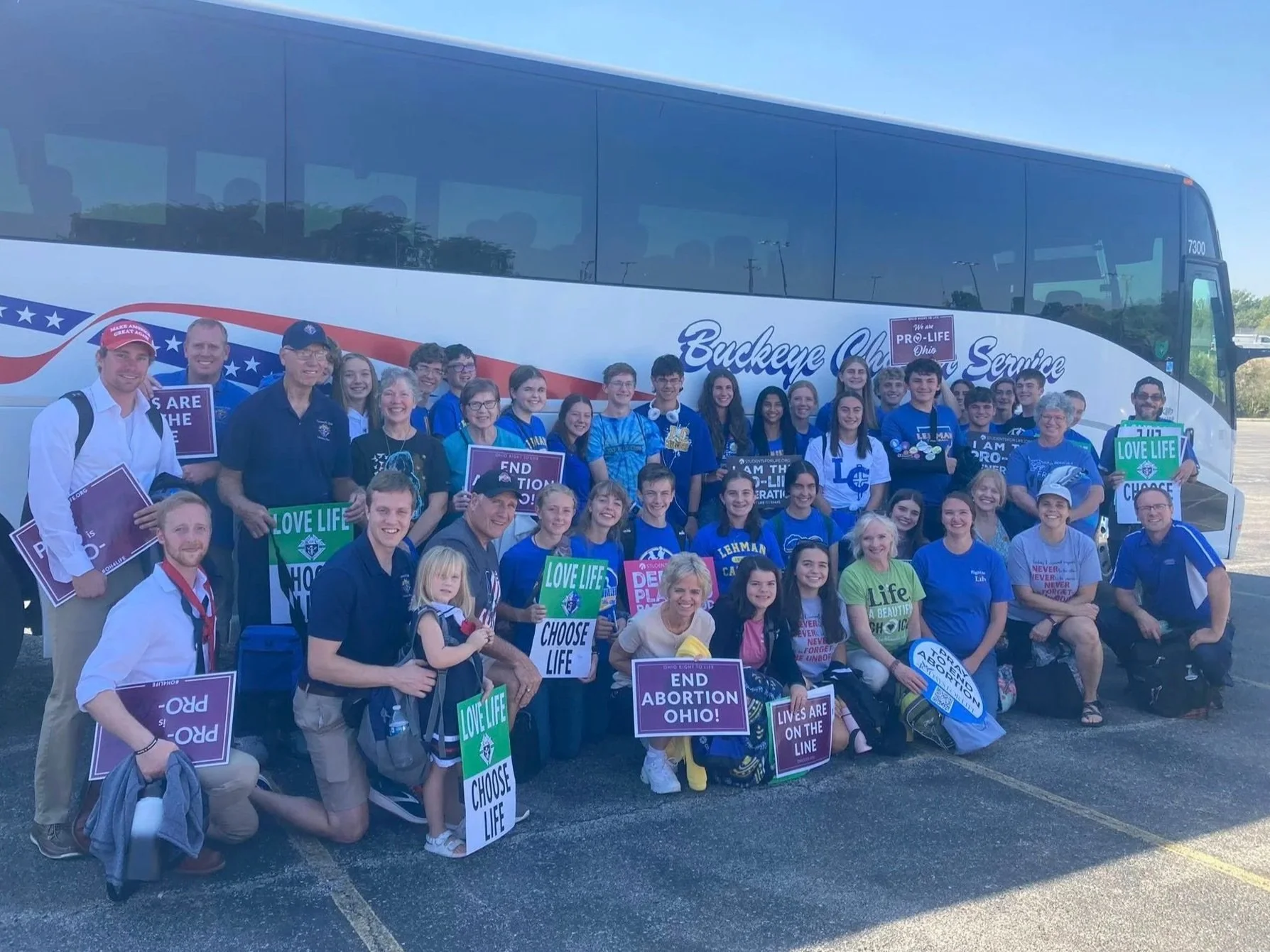 Group of people, including children and adults, standing and kneeling in front of a large bus with Ohio and pro-life signs, holding protest signs supporting pro-life messages.