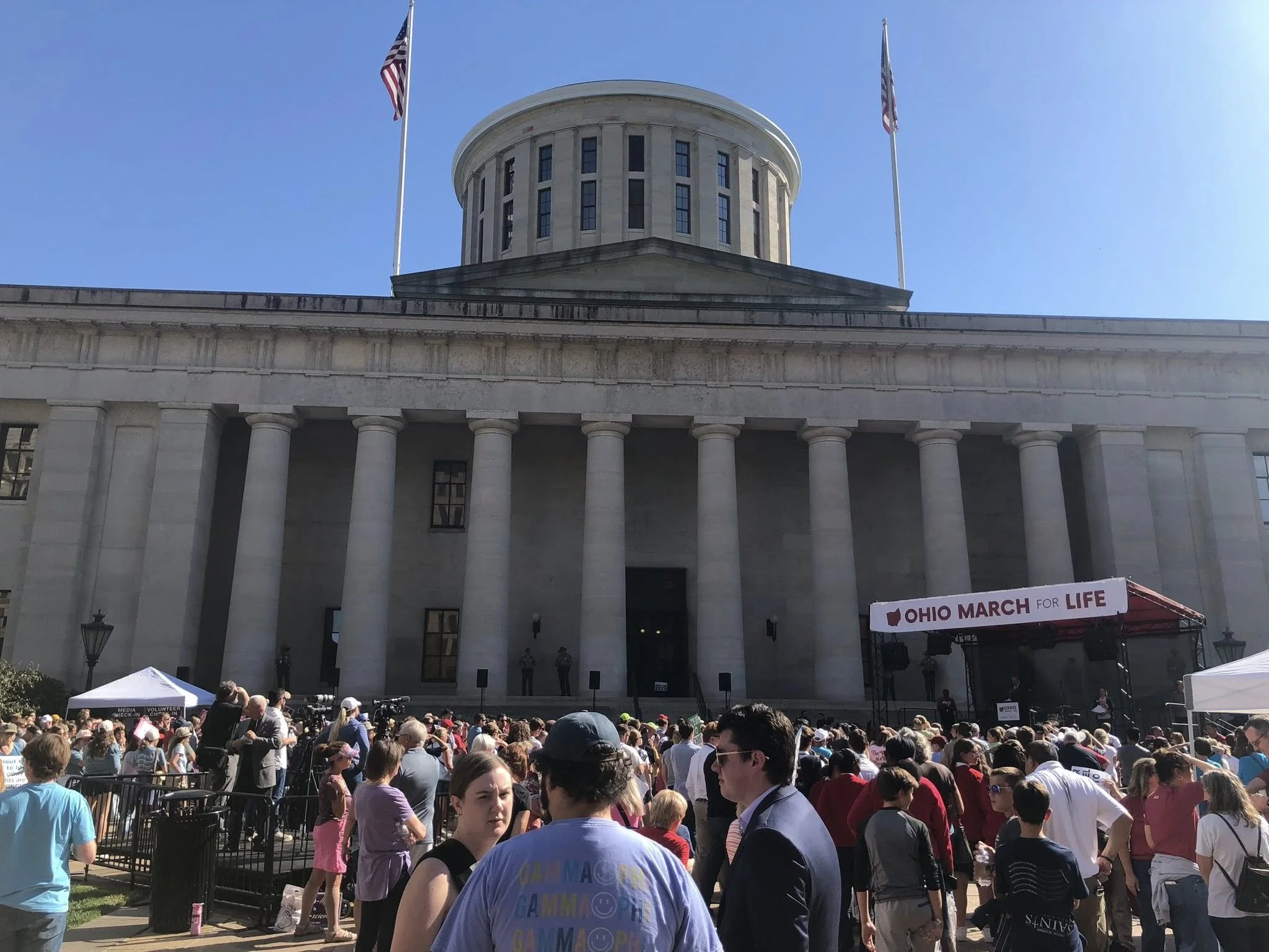 Crowd gathered outside a government building with large columns, watching a stage with a banner that reads 'Ohio March for Life.' The building has a dome and two American flags flying on poles.