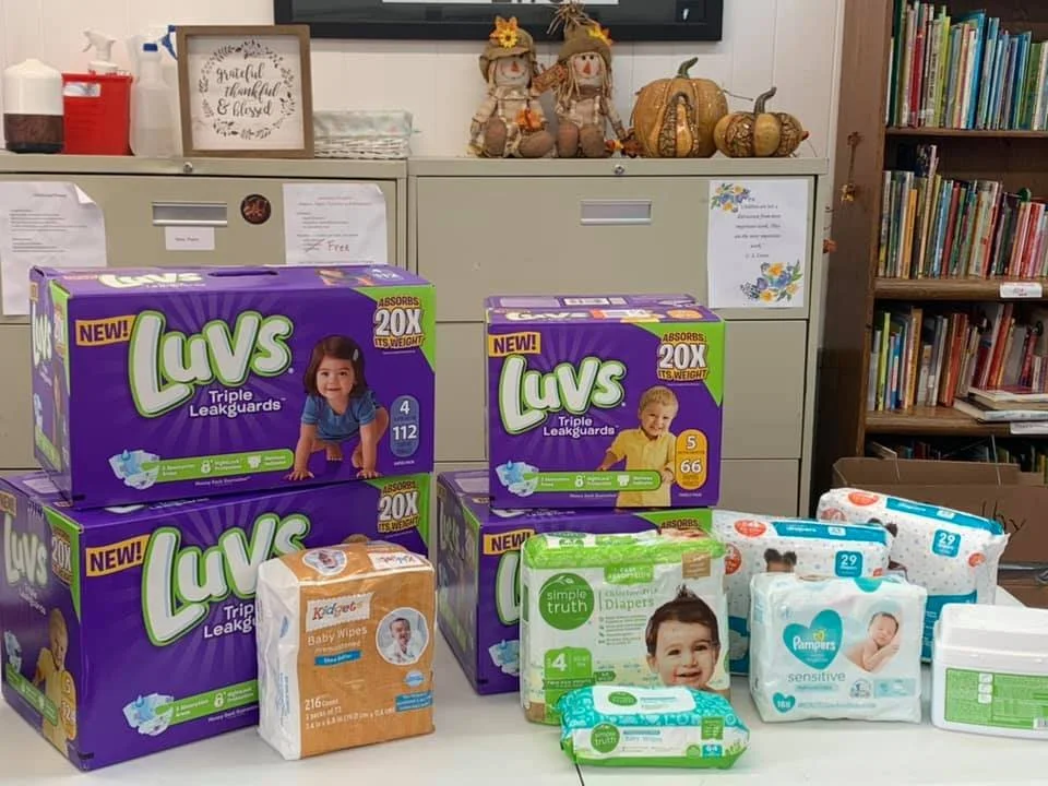 Boxes of Luvs triple leakguard diapers and packages of baby wipes on a table in front of a filing cabinet. In the background, there are autumn-themed decorations including gourds and scarecrows, and a bookshelf filled with books.