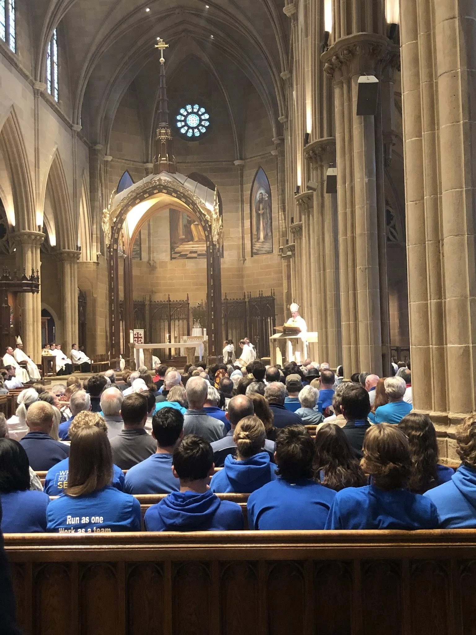 A large congregation inside a grand cathedral, attending a religious service or mass. The interior features high vaulted ceilings, tall columns, religious paintings, and stained glass windows. The clergy are seated at the front, and a speaker is addr