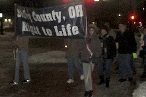 A group of people holding a banner that says 'Shelby County, OH Right to Life' during a night-time demonstration or protest.