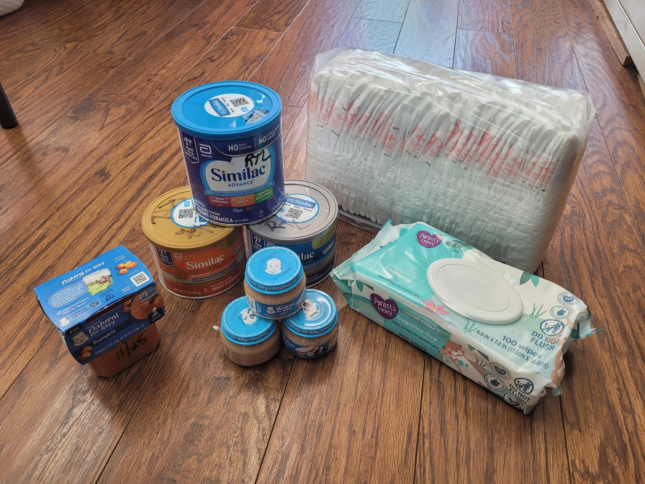 An assortment of baby supplies on a wooden floor, including baby formula, baby wipes, baby food, and a pack of disposable diapers.