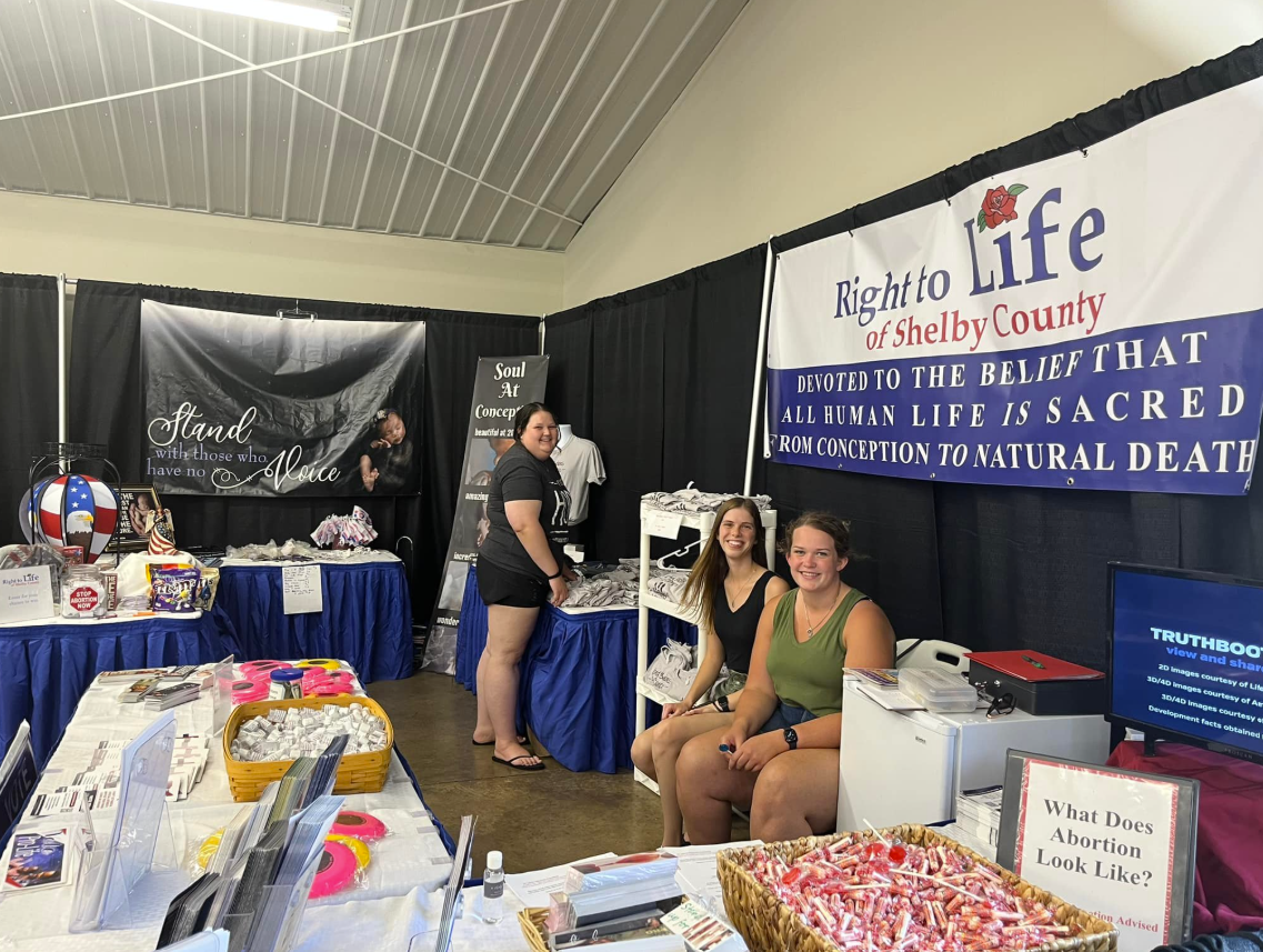 Booth at Shelby County Fair supporting pro-life views with banners, informational materials, and a table of candy. Two women are seated and smiling, while another woman stands behind the table.