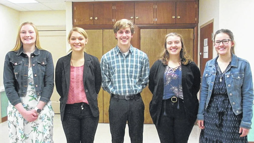 Five young adults standing side by side, smiling, indoors with wooden cabinets behind them.