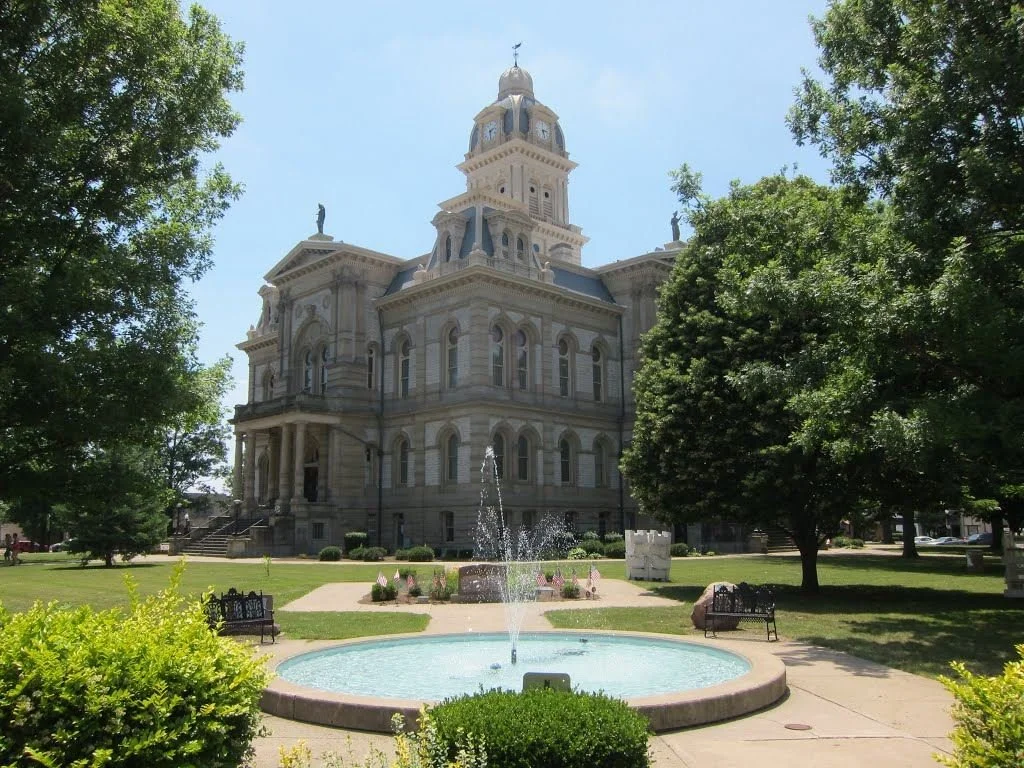 An ornate historic courthouse with a clock tower, surrounded by lush trees and a small fountain in the foreground.