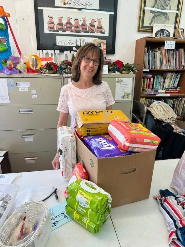 Shelby County Right to Life thrift store in Sidney Ohio; A woman smiling behind a table with a box of supplies, including packs of toilet paper and paper towels, in a room decorated with Christmas decorations and surrounded by bookshelves.