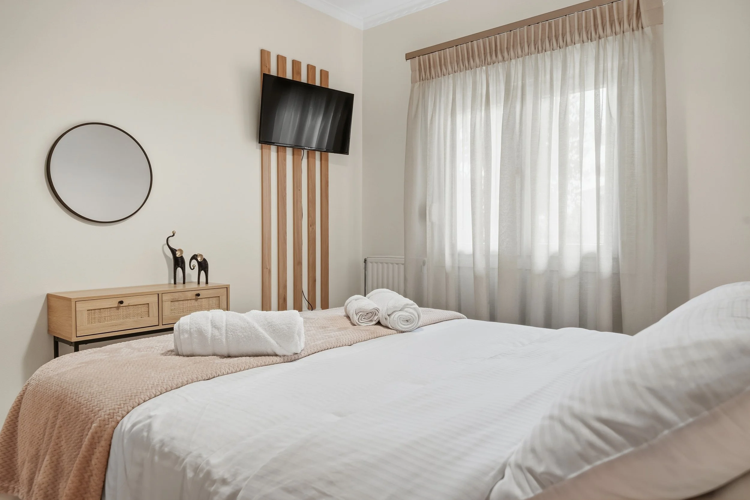 A bedroom with a neatly made bed, white linens, and a beige textured blanket. There are three rolled towels on the bed. On the wall above the bed, there is a small round mirror and a light-colored wooden console table that holds two decorative giraff