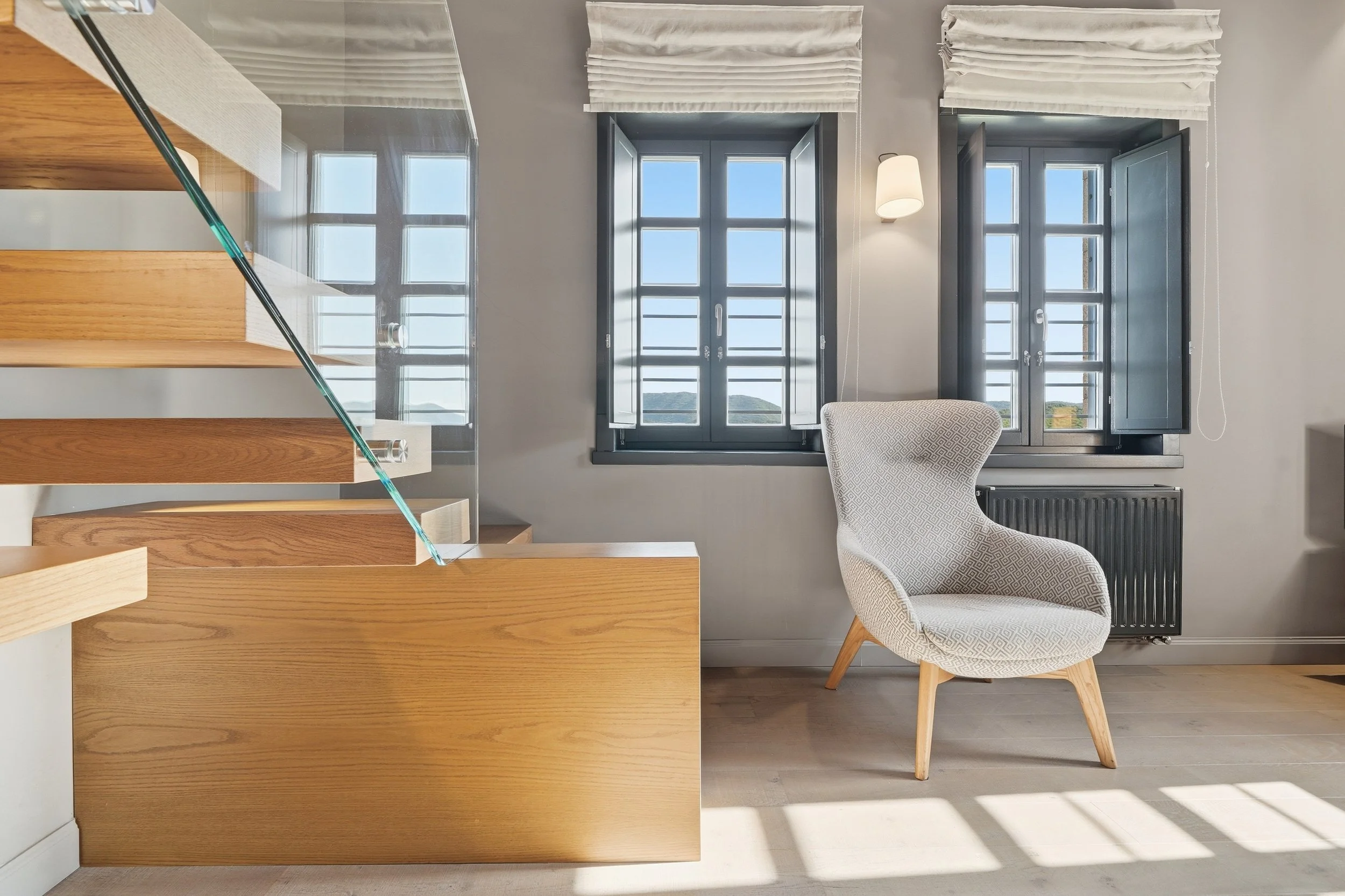 A corner of a room with two windows with blue frames and white curtains, a white wall, a patterned armchair, a black radiator, a hanging light, and wooden staircase steps with a glass railing.