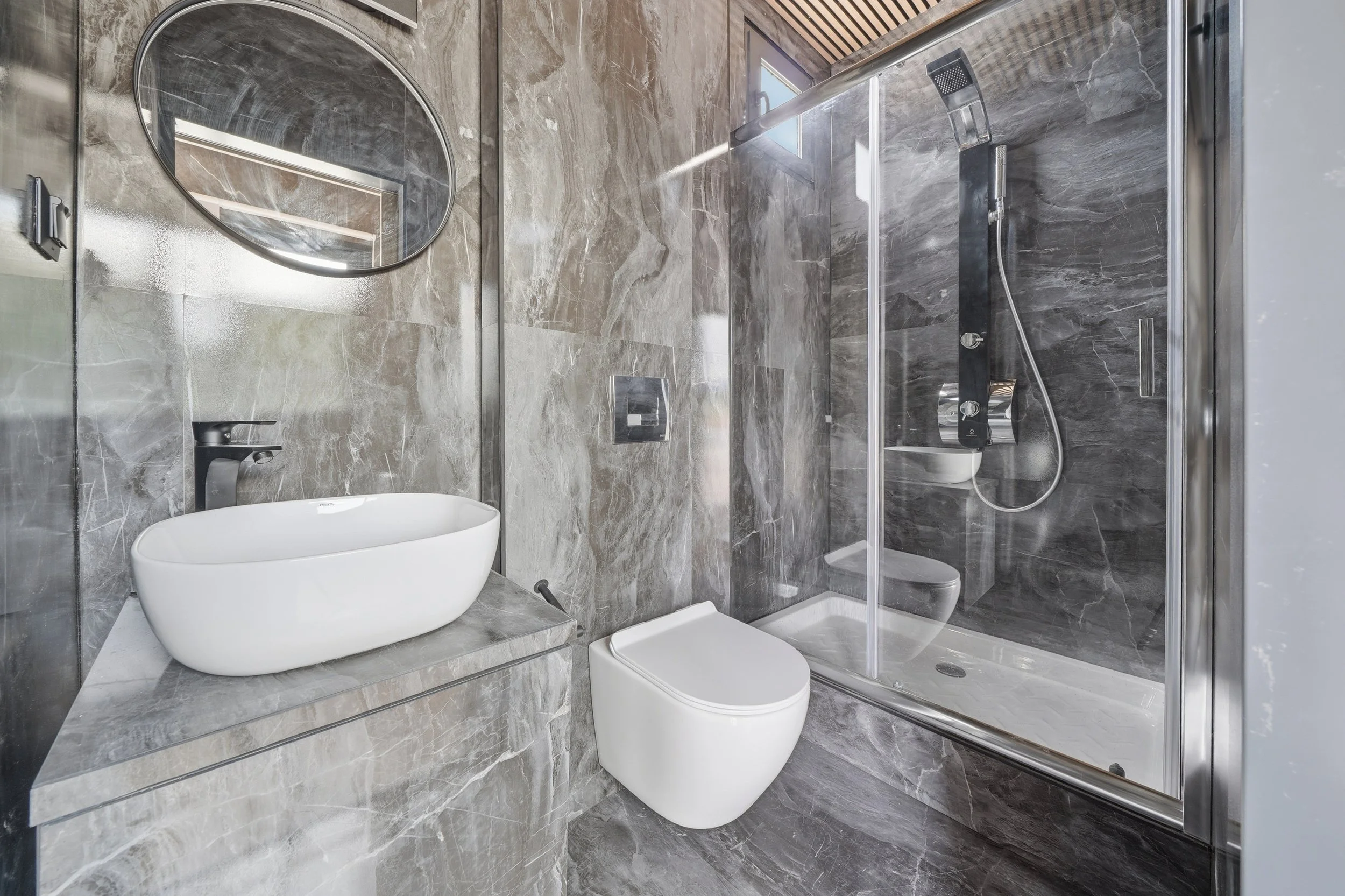 Modern bathroom with grey marble walls and floors, a white oval vessel sink on a grey marble vanity, a wall-mounted toilet, a glass-enclosed shower with a rain showerhead, and a round mirror.