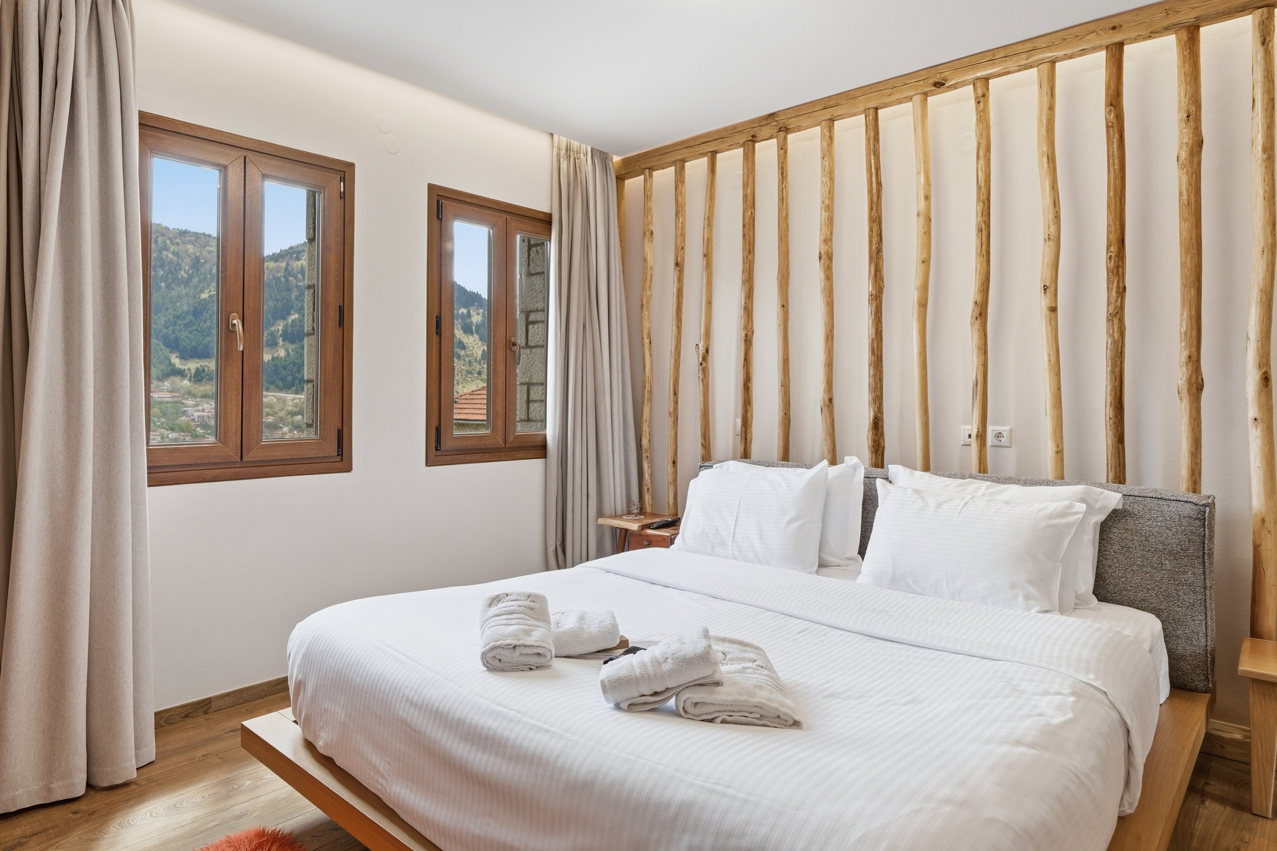 A bedroom with white bedding, towels, and wooden accents, including two windows with curtains showing a mountain view.