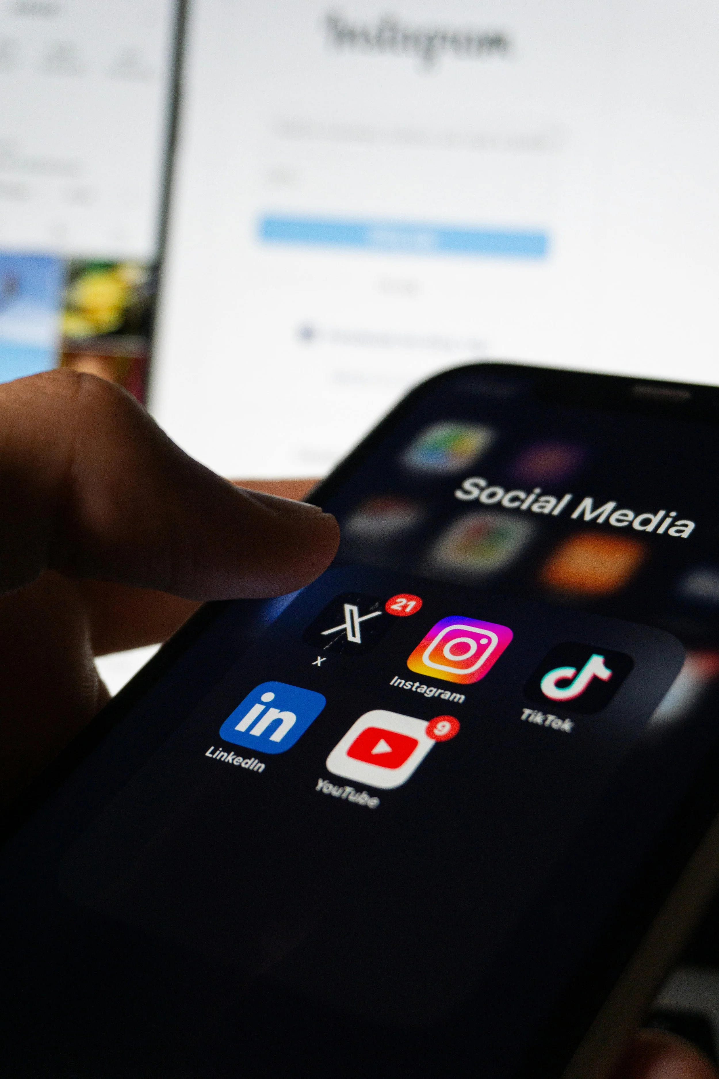 Close-up of a smartphone screen showing social media app icons including Instagram, TikTok, LinkedIn, and YouTube, with a blurred background of a computer monitor displaying a social media application.