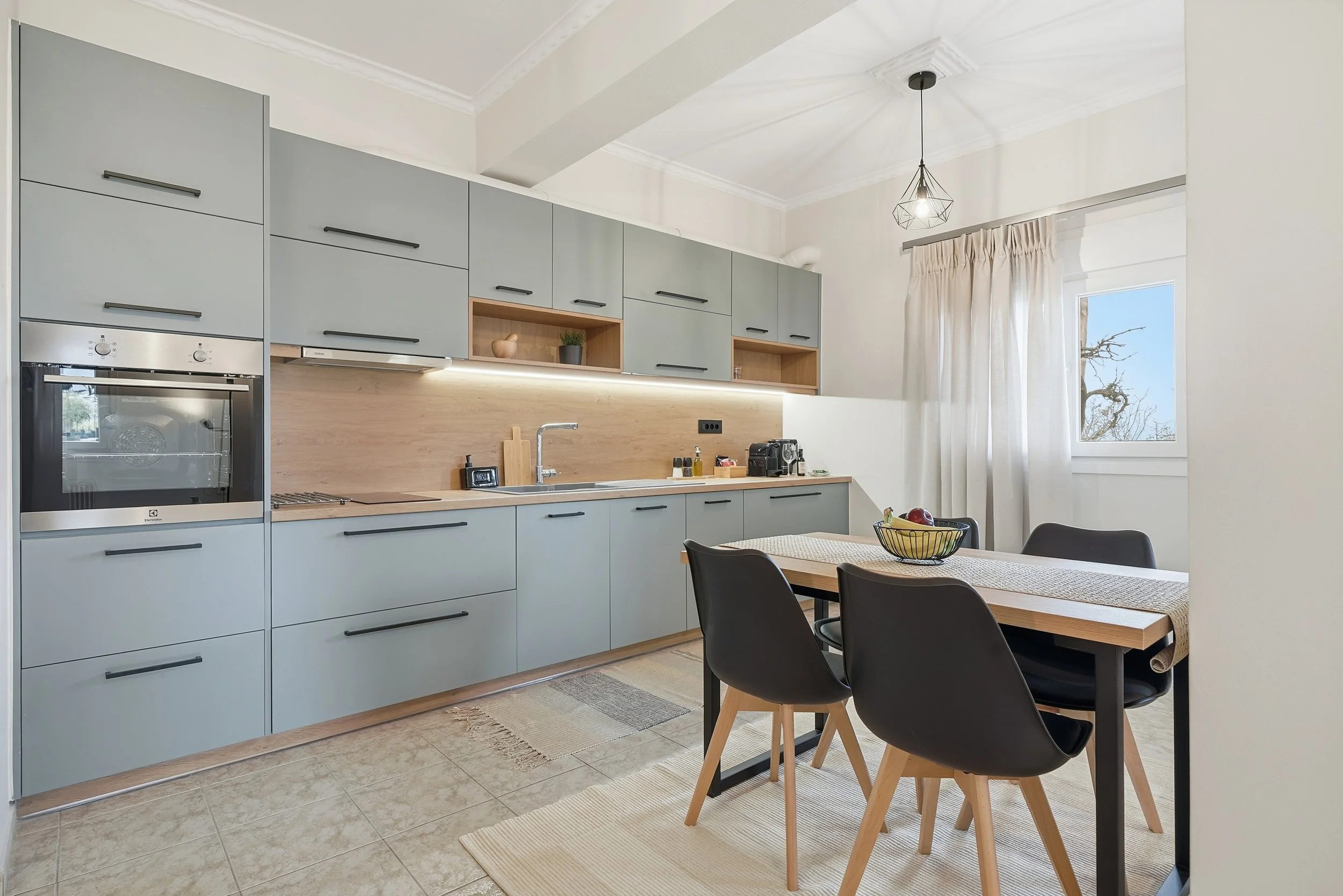 Modern kitchen with light blue cabinets, wooden countertops, and black handles. A dining table with black chairs and a fruit bowl. Window with white curtains, tiled floor, and a ceiling light fixture.