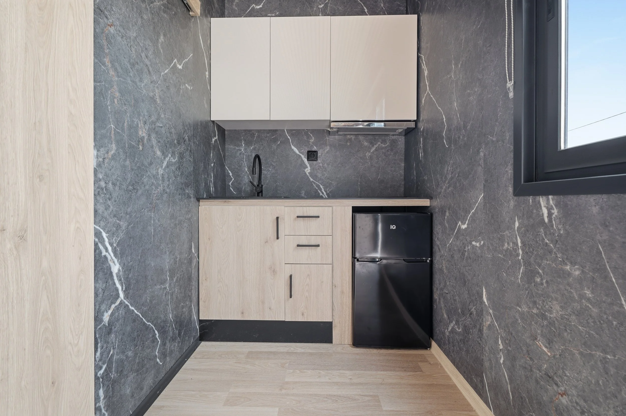 Kitchen with light wood cabinets, a black refrigerator, a black sink with a black faucet, a black electrical outlet on a dark gray marble wall, and a large window with a chain pull cord.