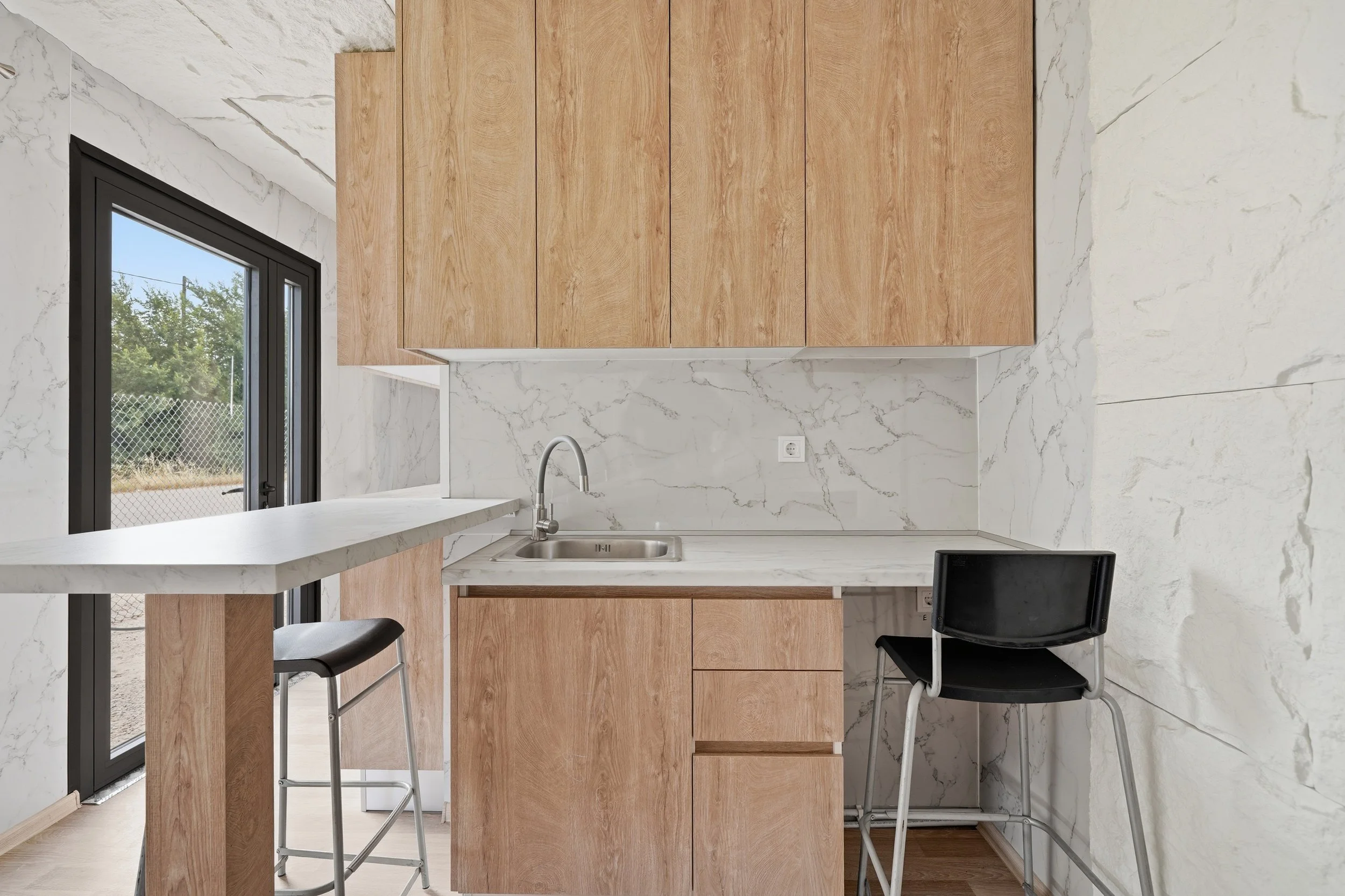 Modern kitchen with white marble walls and a wooden cabinet. There is a black-framed window or door leading outside, a small sink with a faucet, and a black chair near the counter.