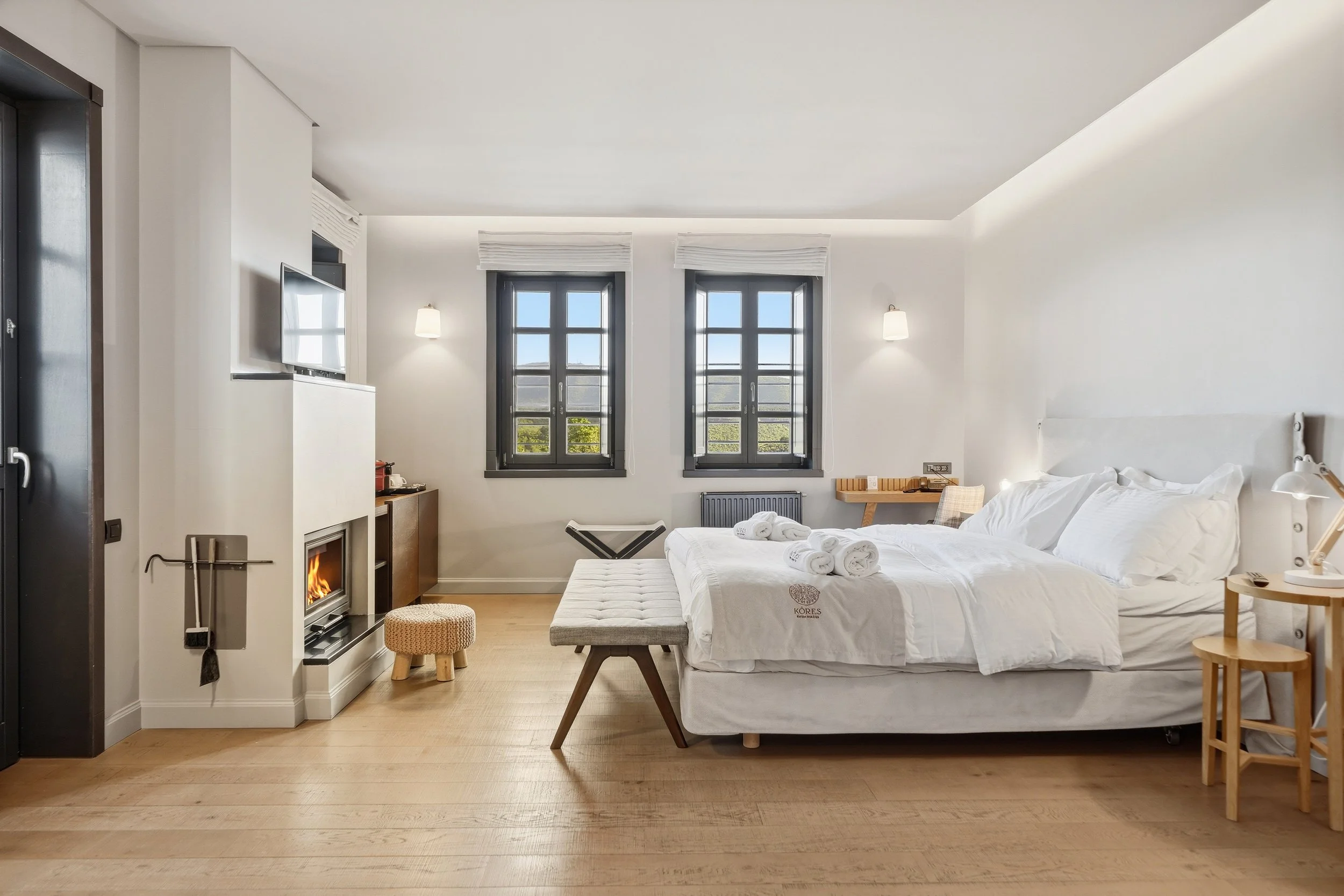 A bright, modern bedroom with two large windows, a white bed with rolled towels, a small ottoman, and a wall-mounted television near a fireplace.